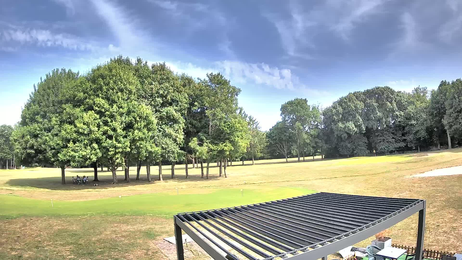 A partly sunny day reveals a golf course with a group of people sitting under trees near a modern pergola.