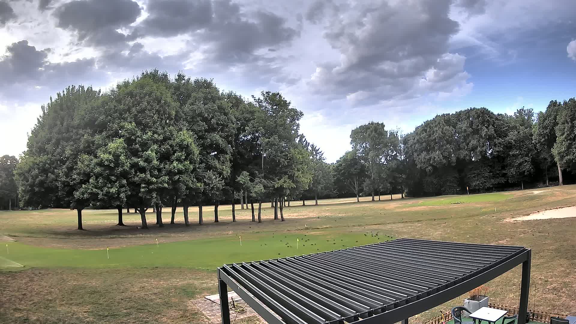 A mostly cloudy sky hangs over a grassy golf course with several trees and a slatted pergola.