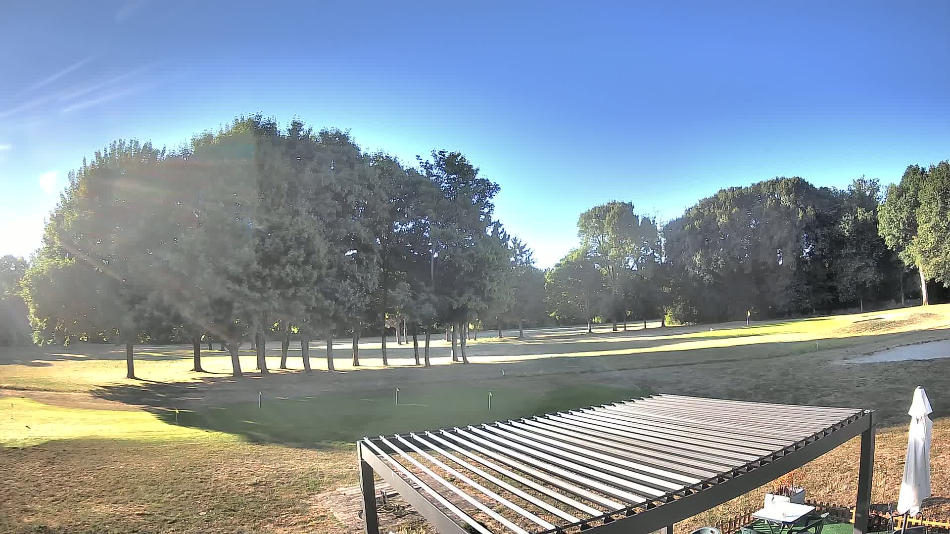 A sunny day on a golf course with a line of trees in the middle ground and a pergola in the foreground.