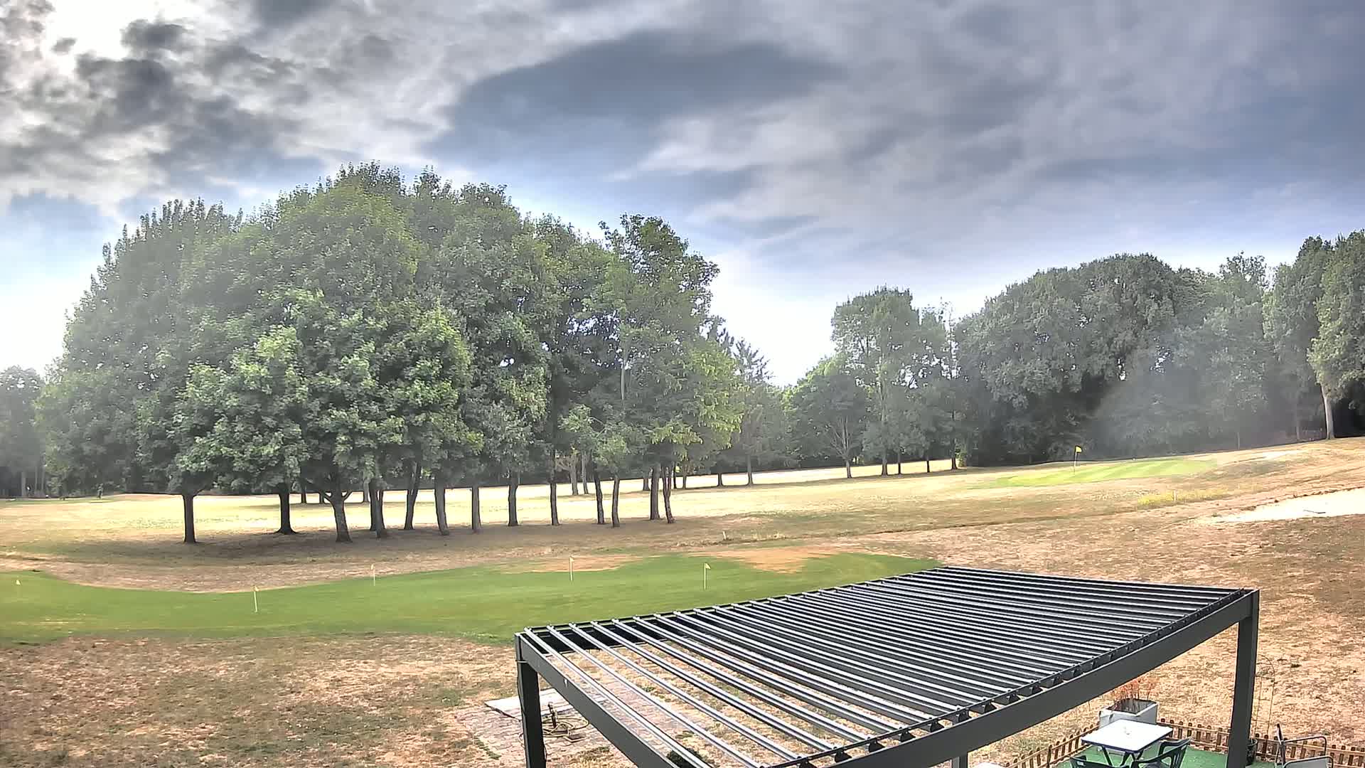 A mostly sunny golf course with a metal pergola in the foreground, under a partly cloudy sky.