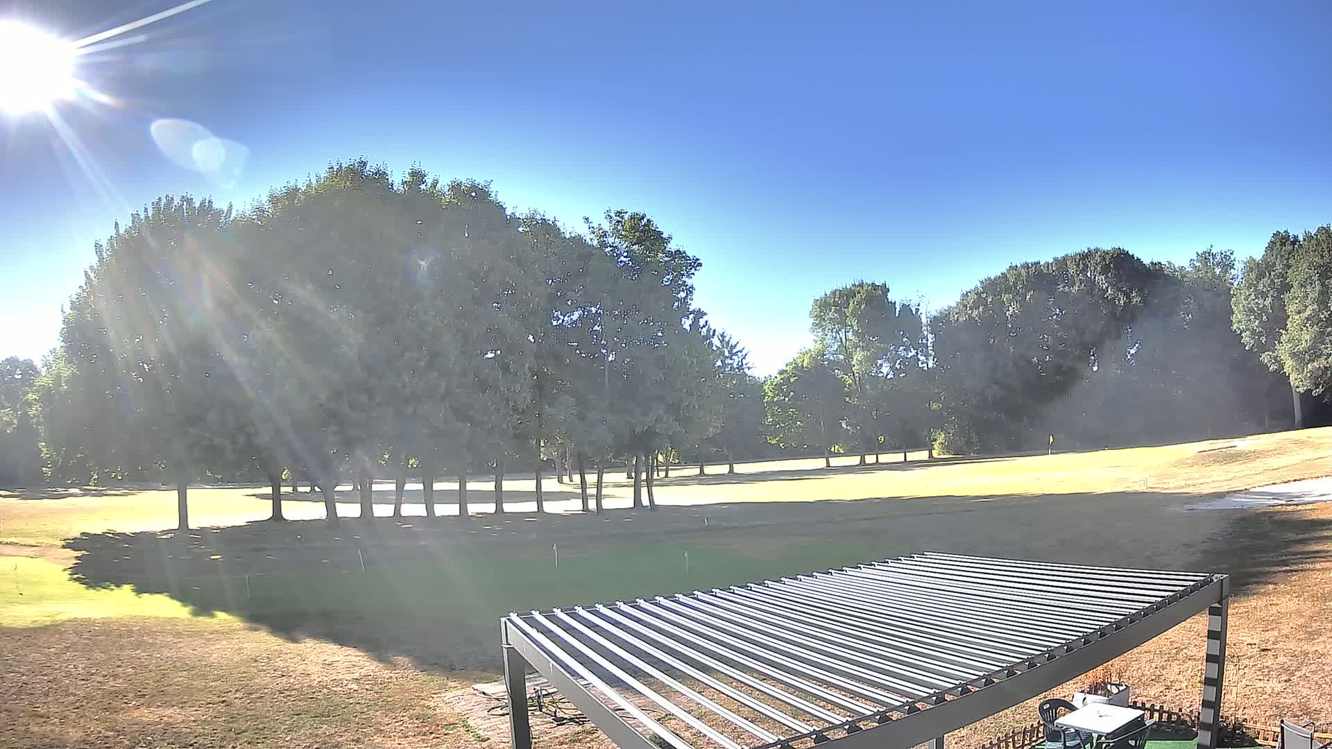 A sunny day on a golf course, viewed from near a pergola, shows a putting green surrounded by trees.