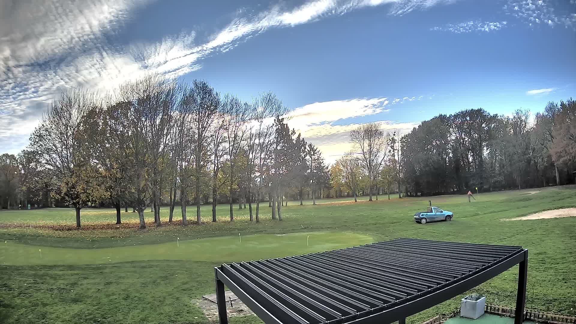 A vibrant golf course featuring a putting green with flags, numerous trees in various stages of leaf-fall, a person golfing in the distance, and a light blue utility vehicle is depicted under a bright, partly cloudy sky, with a dark slatted pergola roof framing the bottom foreground.