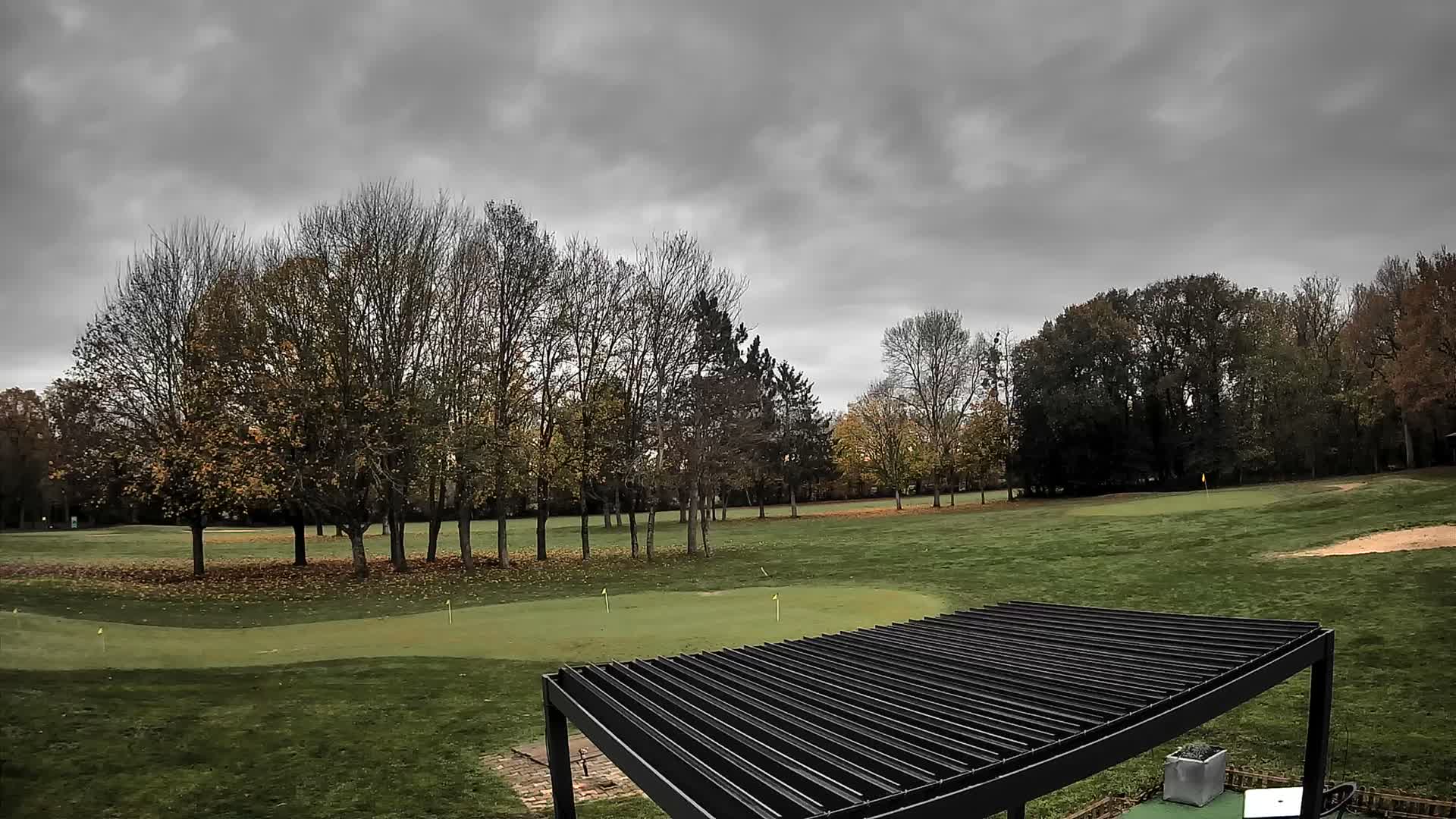 A vibrant golf course featuring a putting green with flags, numerous trees in various stages of leaf-fall, a person golfing in the distance, and a light blue utility vehicle is depicted under a bright, partly cloudy sky, with a dark slatted pergola roof framing the bottom foreground.