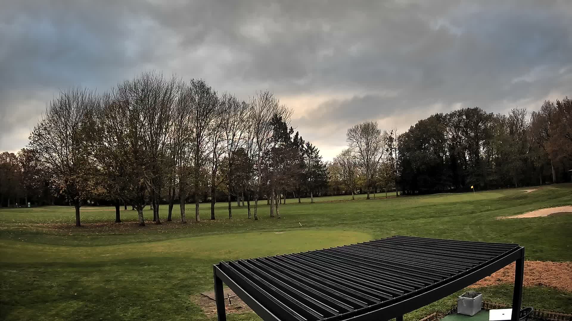 Under a heavily overcast and dark sky, a golf course with green fairways, a putting green featuring a flag, scattered trees (many bare), and a sand trap is partially observed behind a black slatted pergola in the foreground.