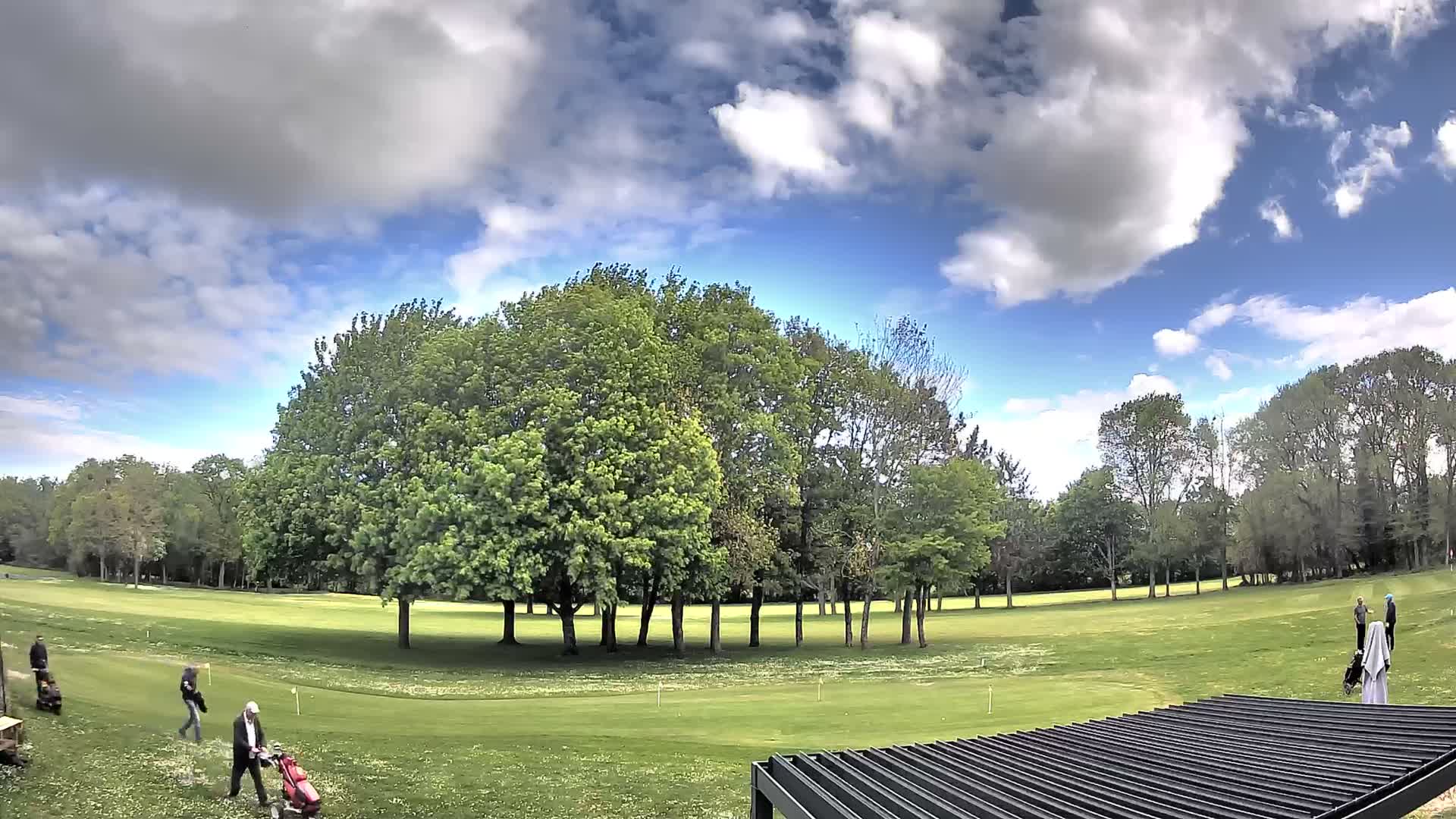 Under a heavily overcast and dark sky, a golf course with green fairways, a putting green featuring a flag, scattered trees (many bare), and a sand trap is partially observed behind a black slatted pergola in the foreground.