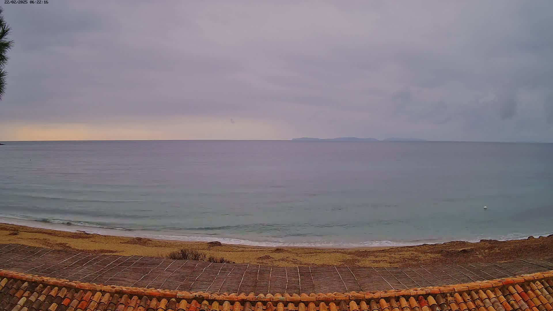 A sandy beach with gentle waves meets a calm sea under an overcast sky, viewed from a tiled roof overlooking a woven mat structure.