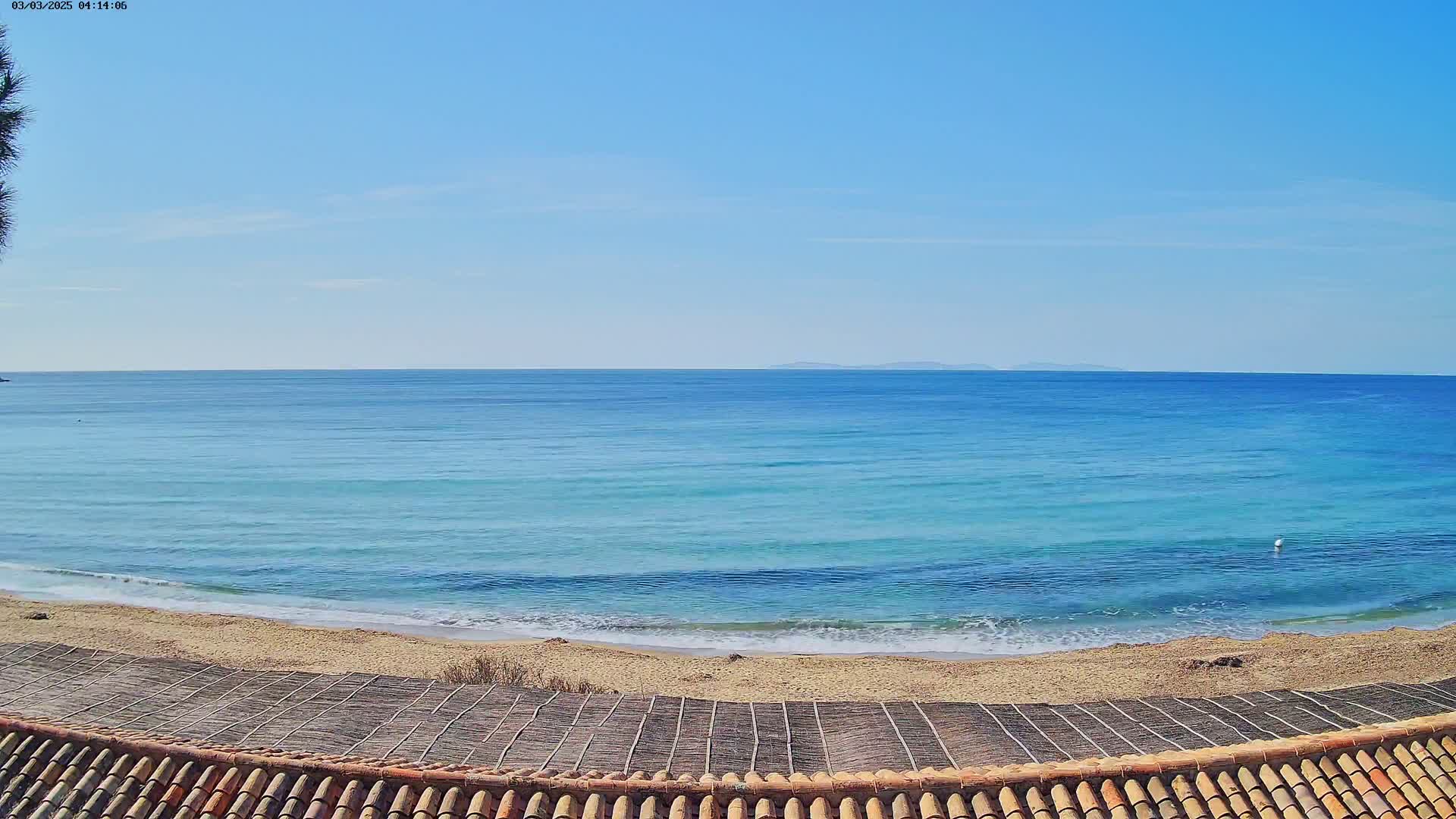 A sandy beach with gentle waves and a calm, clear blue ocean under a bright, sunny sky is viewed from behind a tiled roof.