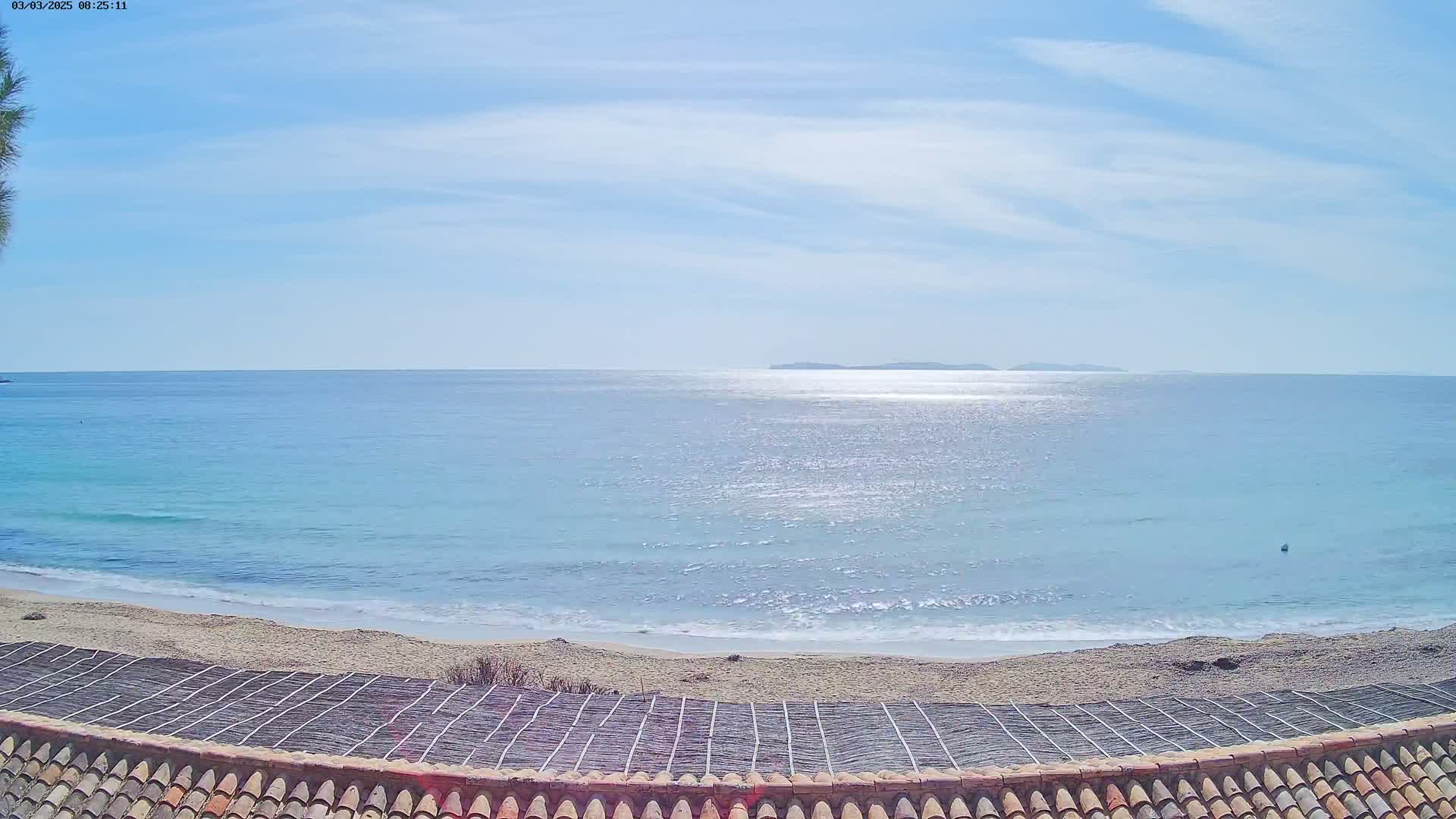 A sandy beach with calm, light-blue water and distant islands under a mostly clear, sunny sky is viewed from atop a tiled roof.