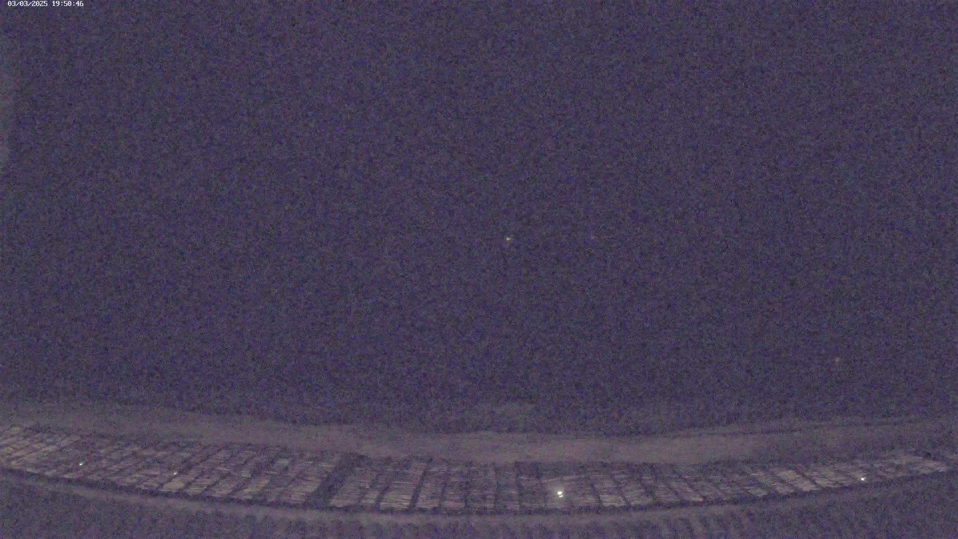 A nighttime, low-light view of a large, flat expanse of land with a grid-like pattern, possibly a field or parking lot, under a dark, mostly clear sky.