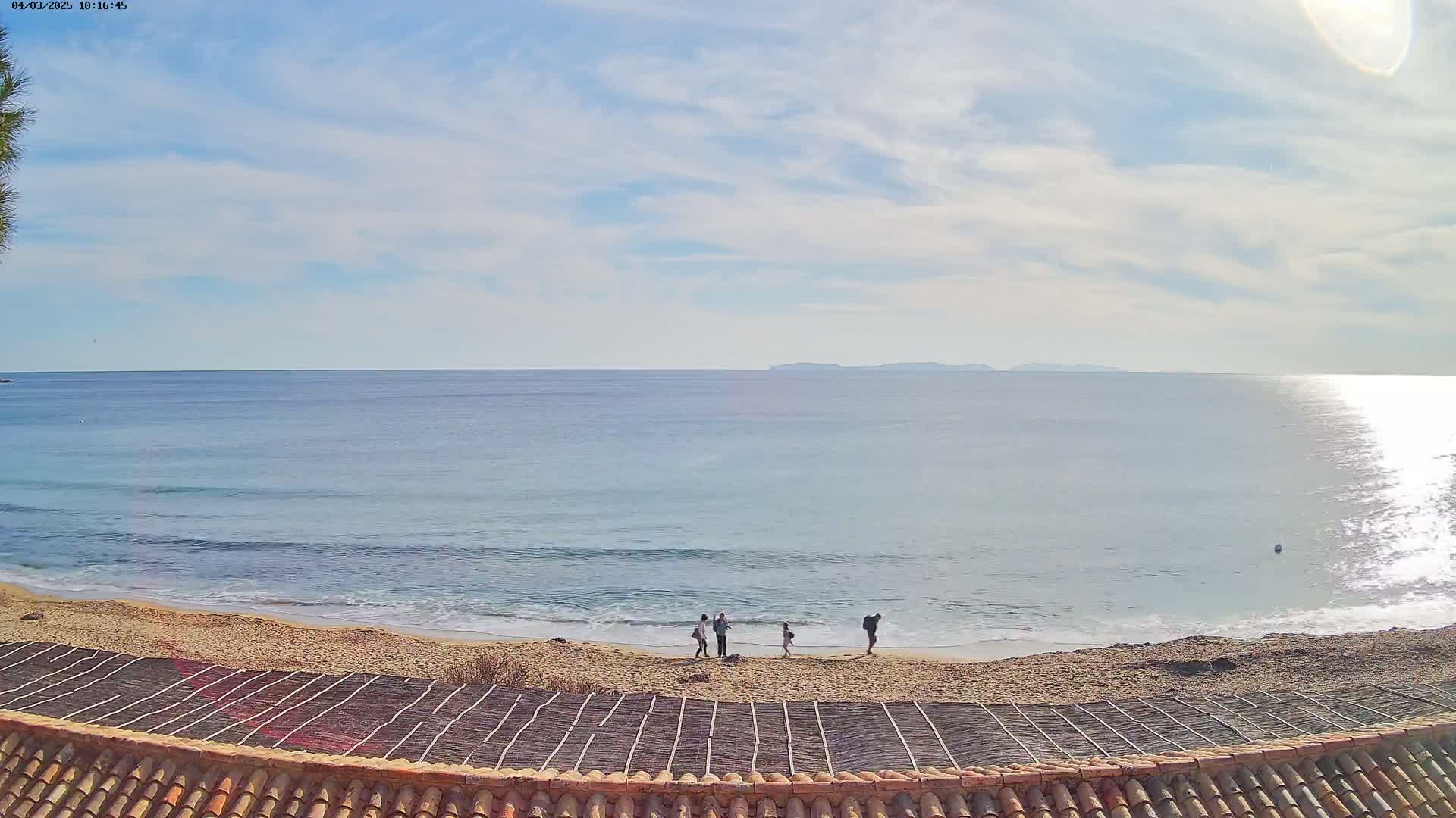 A sandy beach with a few people on it is visible under a partly cloudy sky, with calm ocean waves lapping the shore and islands in the distance.