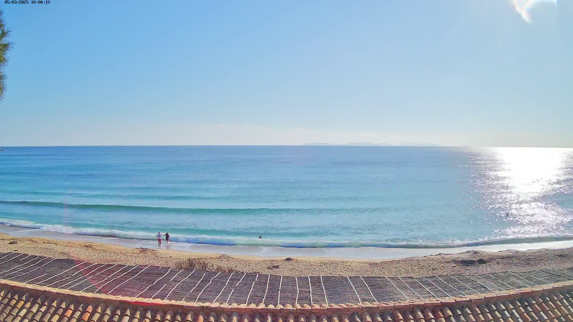 A sandy beach with gentle waves and a few people, under a clear sunny sky, is viewed from above a tiled roof.