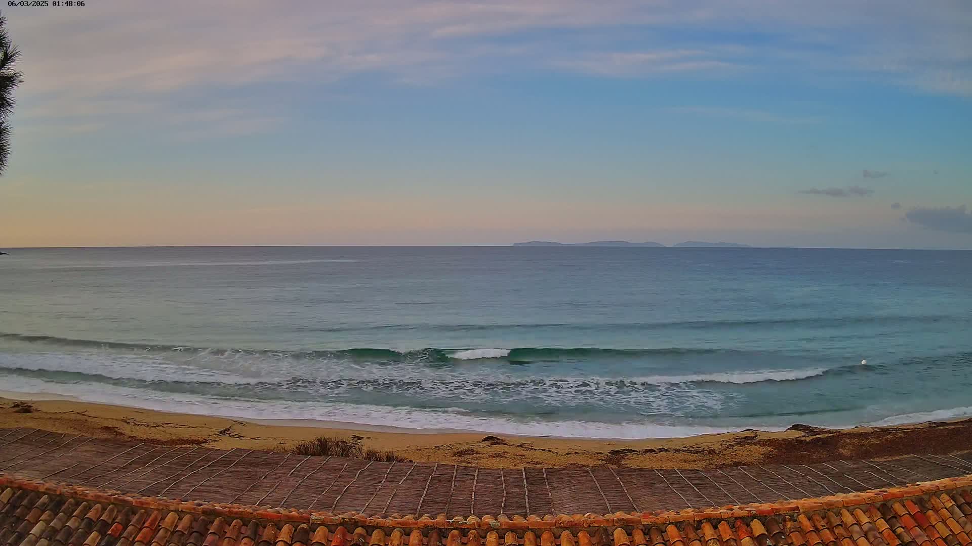 Gentle waves roll onto a sandy beach under a mostly clear, pale sky, with islands visible on the horizon.
