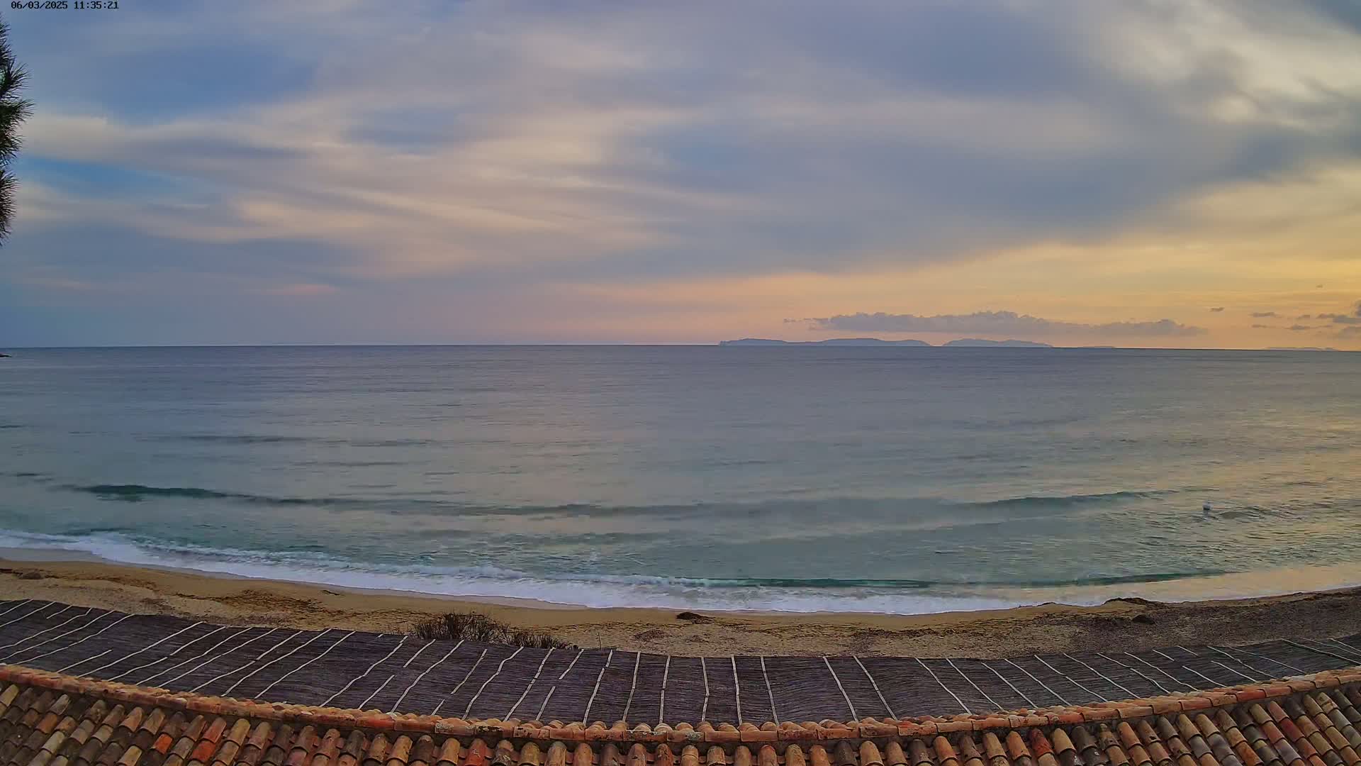 A sandy beach with gentle waves laps the shore under a partly cloudy sunset sky, with an island visible on the horizon.