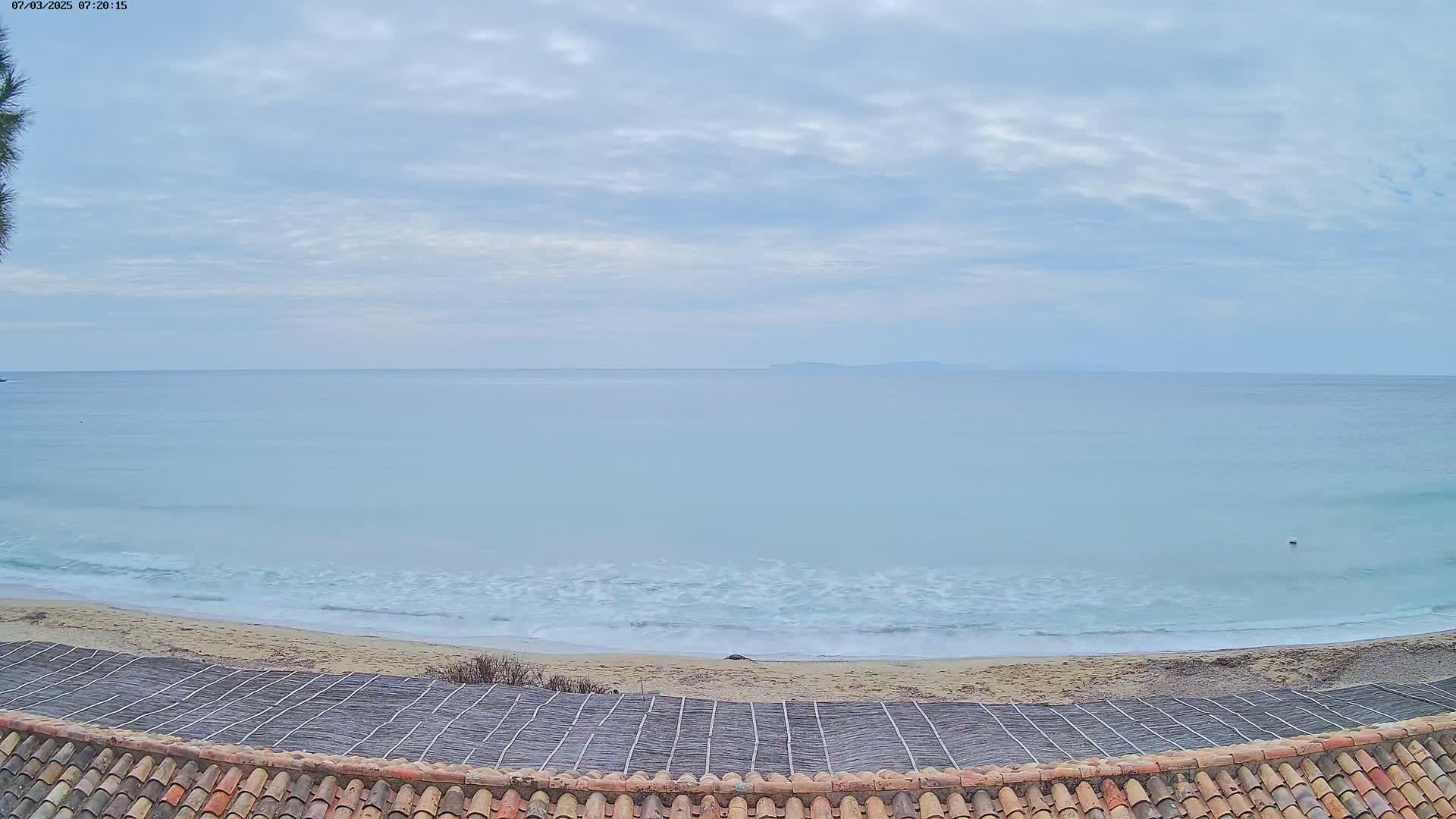 A sandy beach with gentle waves under a cloudy sky meets a calm ocean with a distant landmass visible on the horizon.
