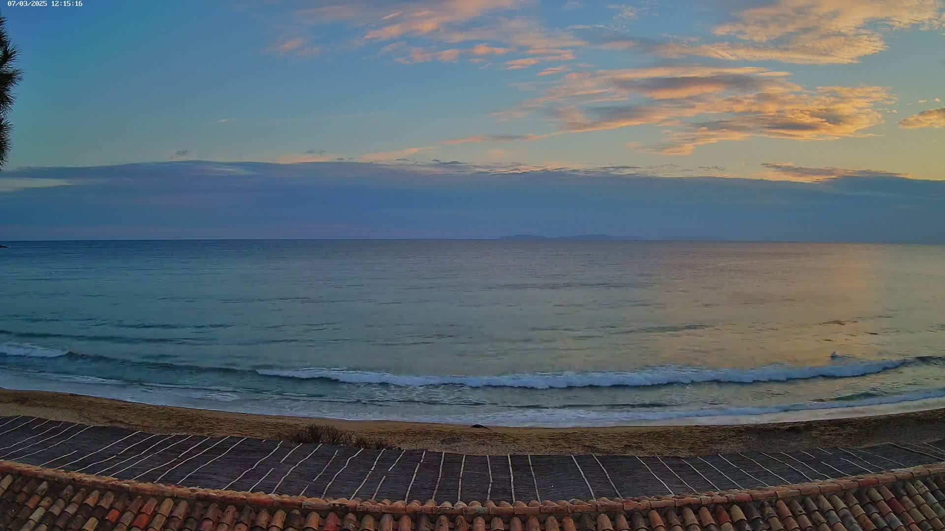 A calm ocean with small waves laps a sandy beach under a partly cloudy sky with hints of sunset.