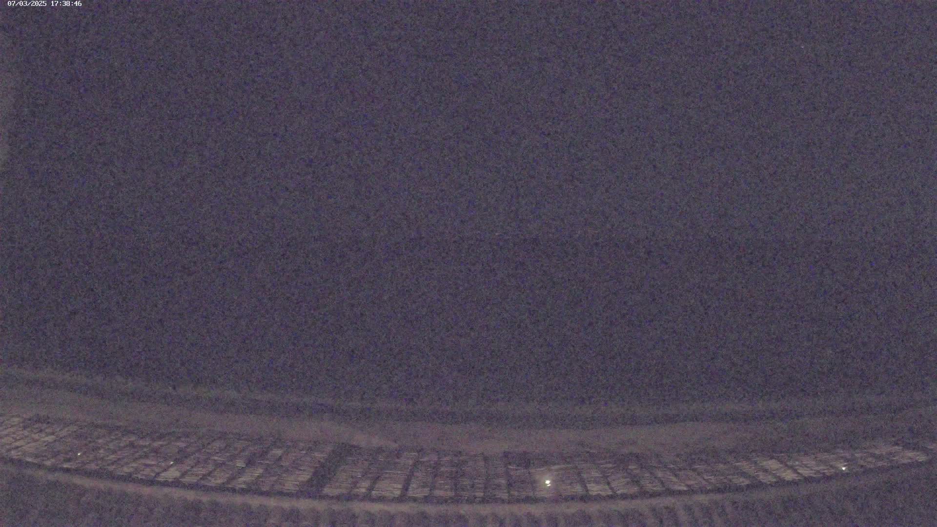 A nighttime, low-light view of a large, flat area, possibly a field or parking lot, with faint illumination visible in the lower portion of the image.