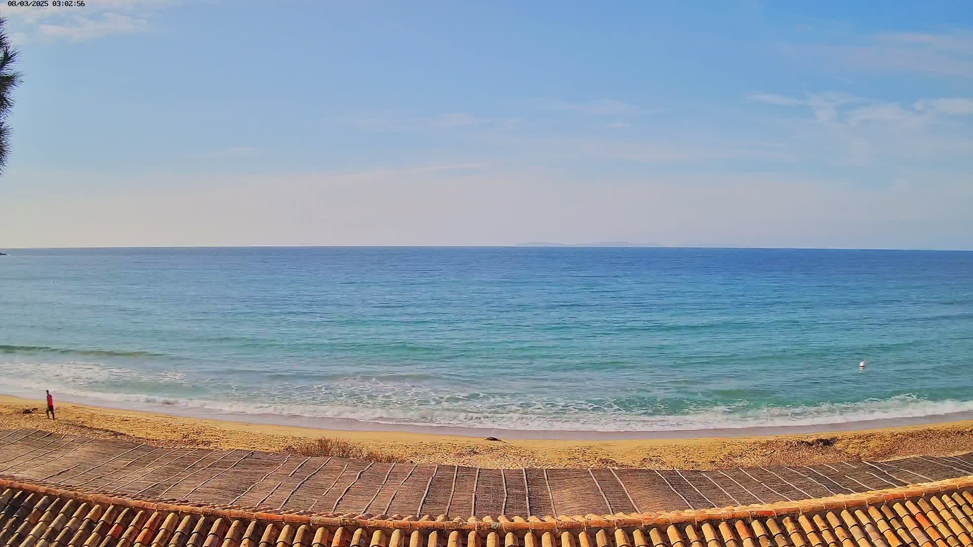 A sandy beach with gentle waves laps the shore under a clear, sunny sky, viewed from a rooftop with a tiled edge.