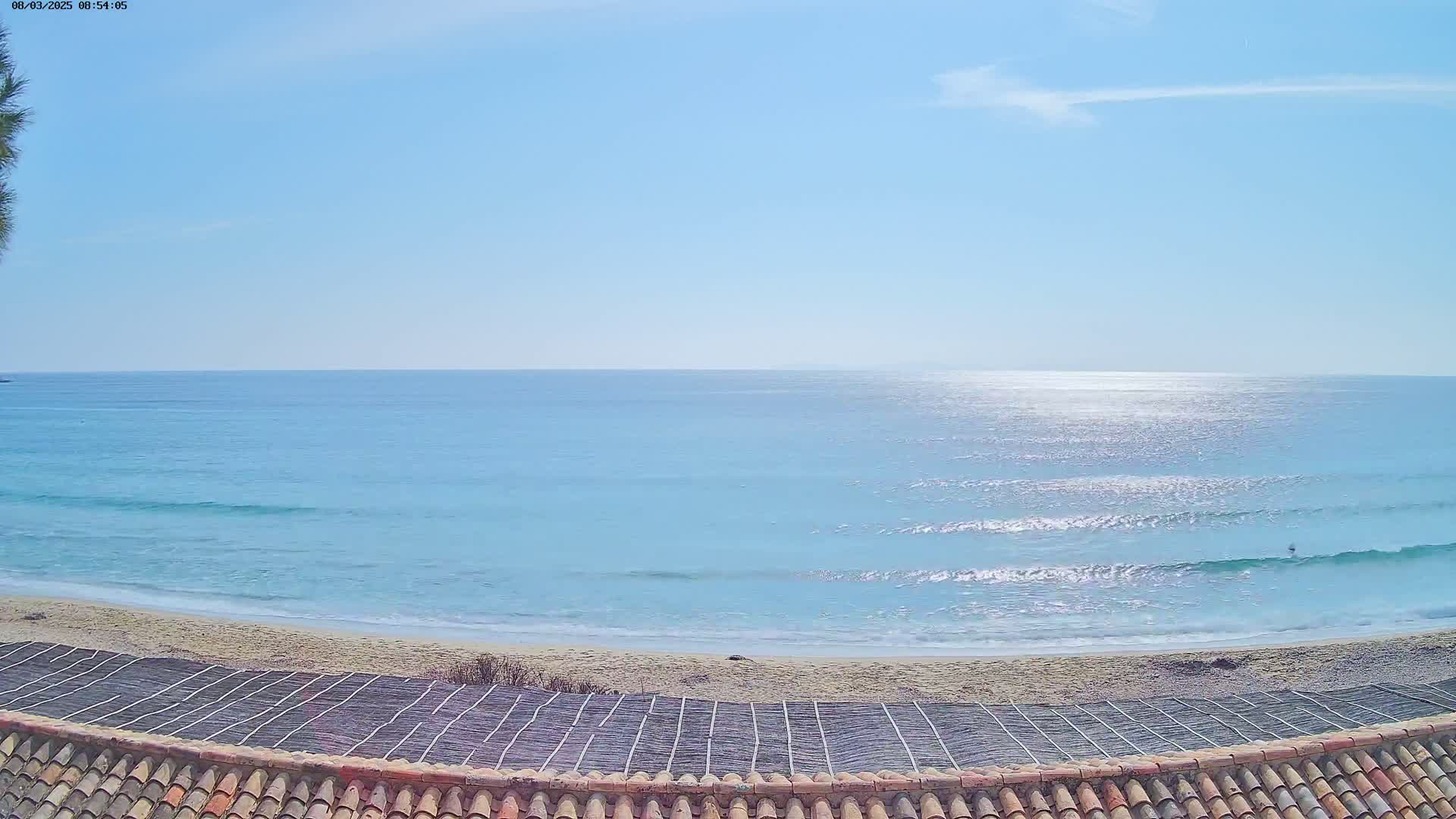 A sandy beach with gentle waves laps at the shore under a clear blue sky, viewed from behind a tiled roof.