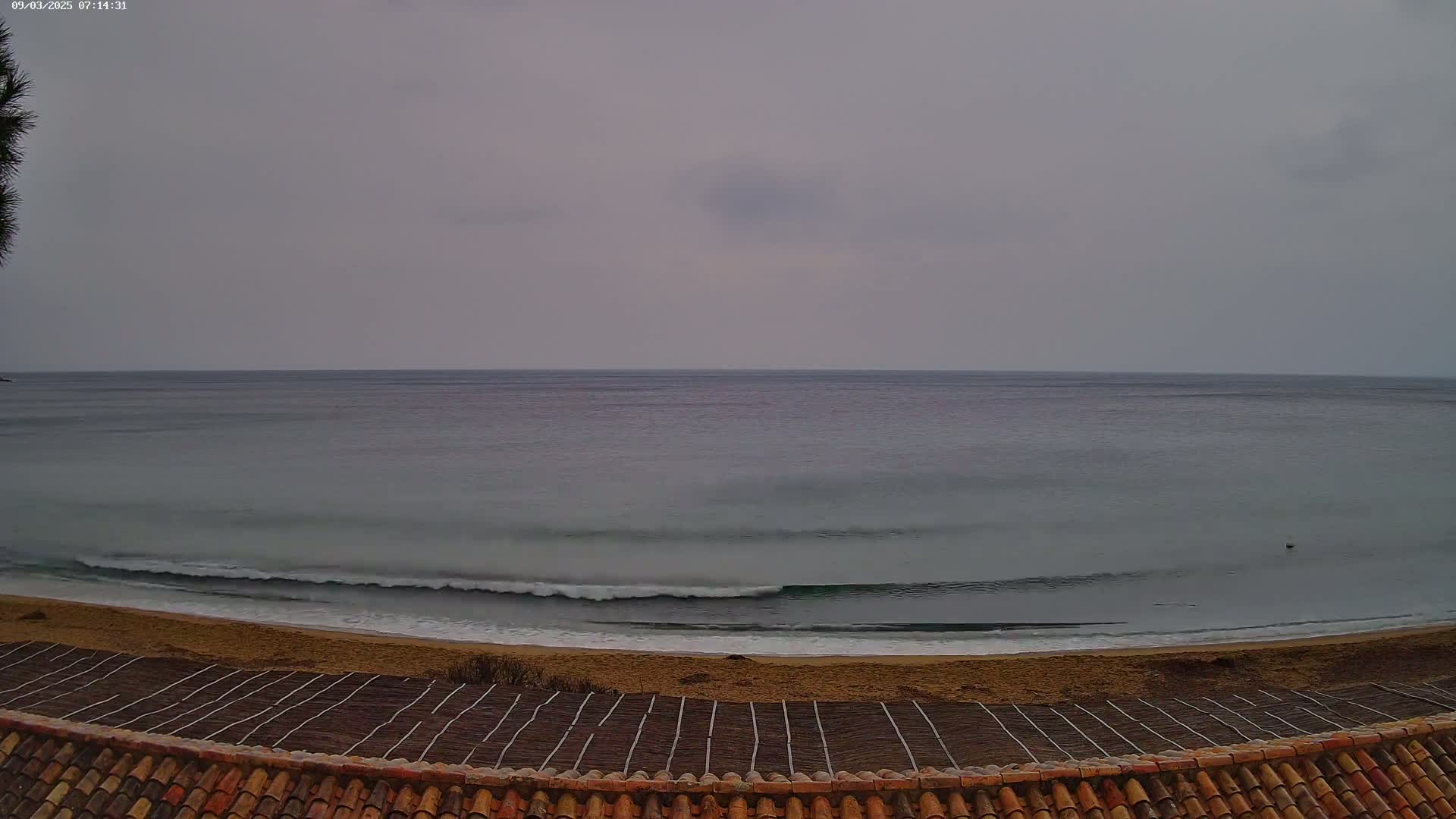 A calm sea with small waves laps a sandy beach, viewed from behind a terracotta roof under an overcast sky.