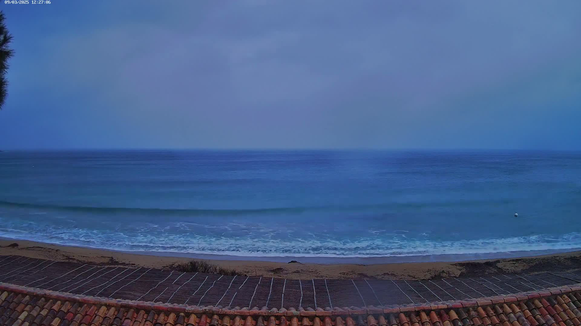 A view from a tiled roof shows a calm, blue ocean meeting a sandy beach under an overcast sky.