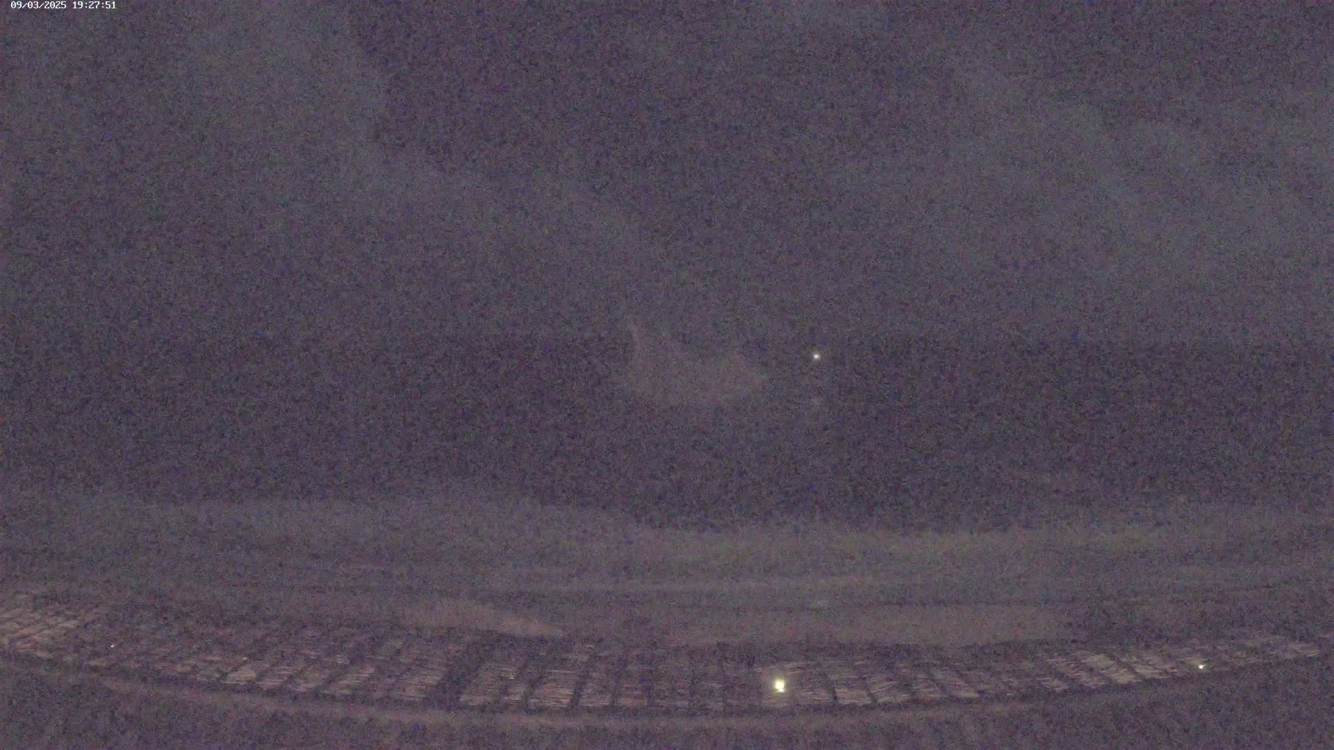 A dark, nighttime scene shows a faintly lit, gridded ground area beneath a mostly cloudy sky with a few stars visible.