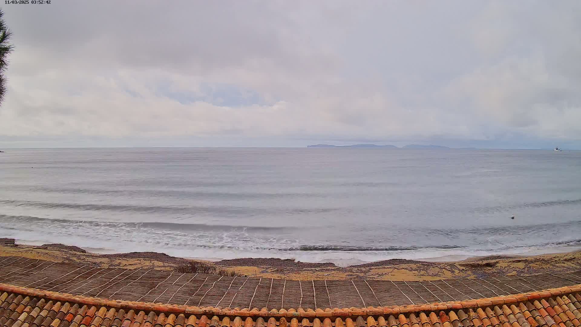 A wavy ocean under a cloudy sky is viewed from behind a low, reddish-brown tiled roof and a section of woven matting.