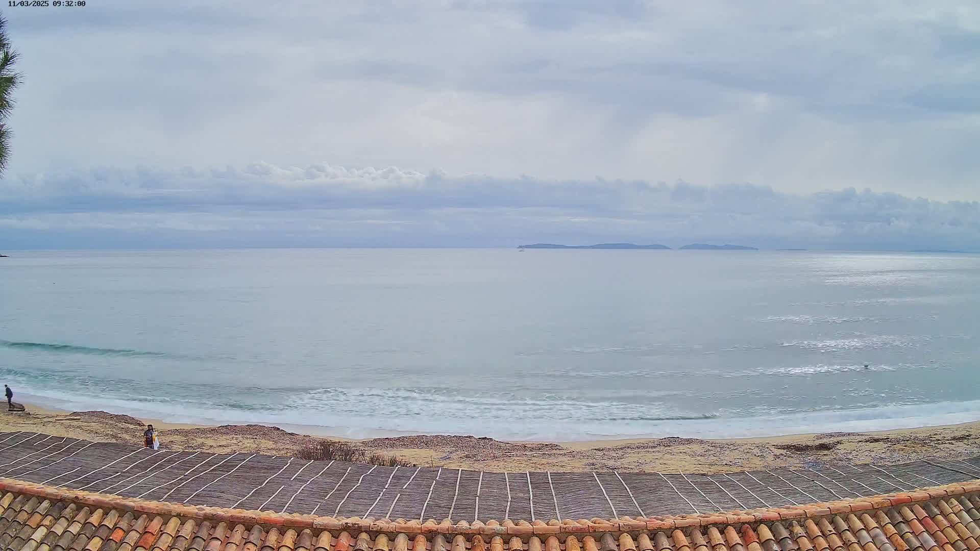 A view from a tiled roof shows a calm ocean with small waves lapping a sandy beach under a cloudy sky, and a few people are visible on the beach.