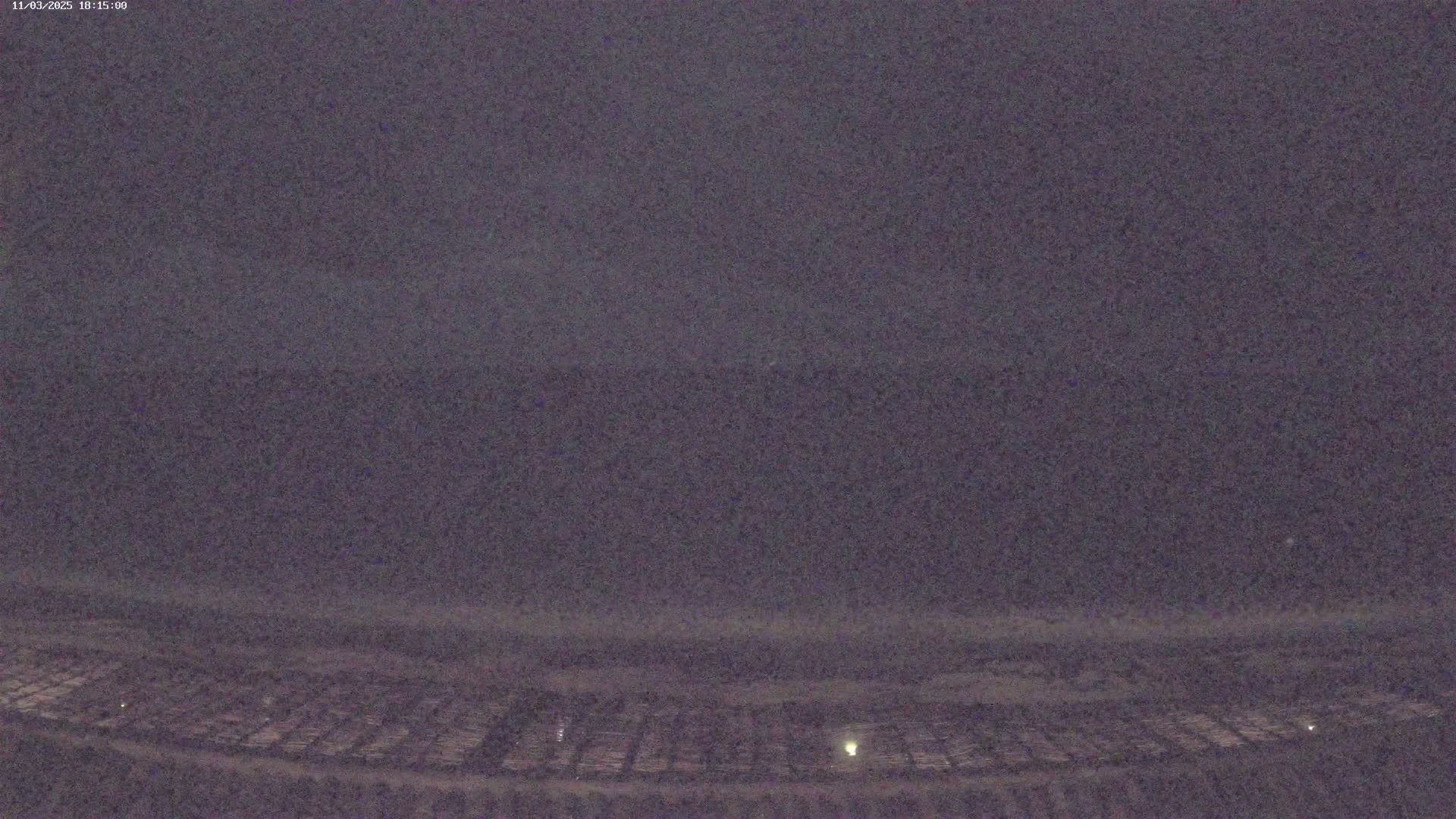 A dark, nighttime view shows a flat expanse of land with faint, regularly spaced lights, extending to a dark horizon under a mostly clear, dusky sky.