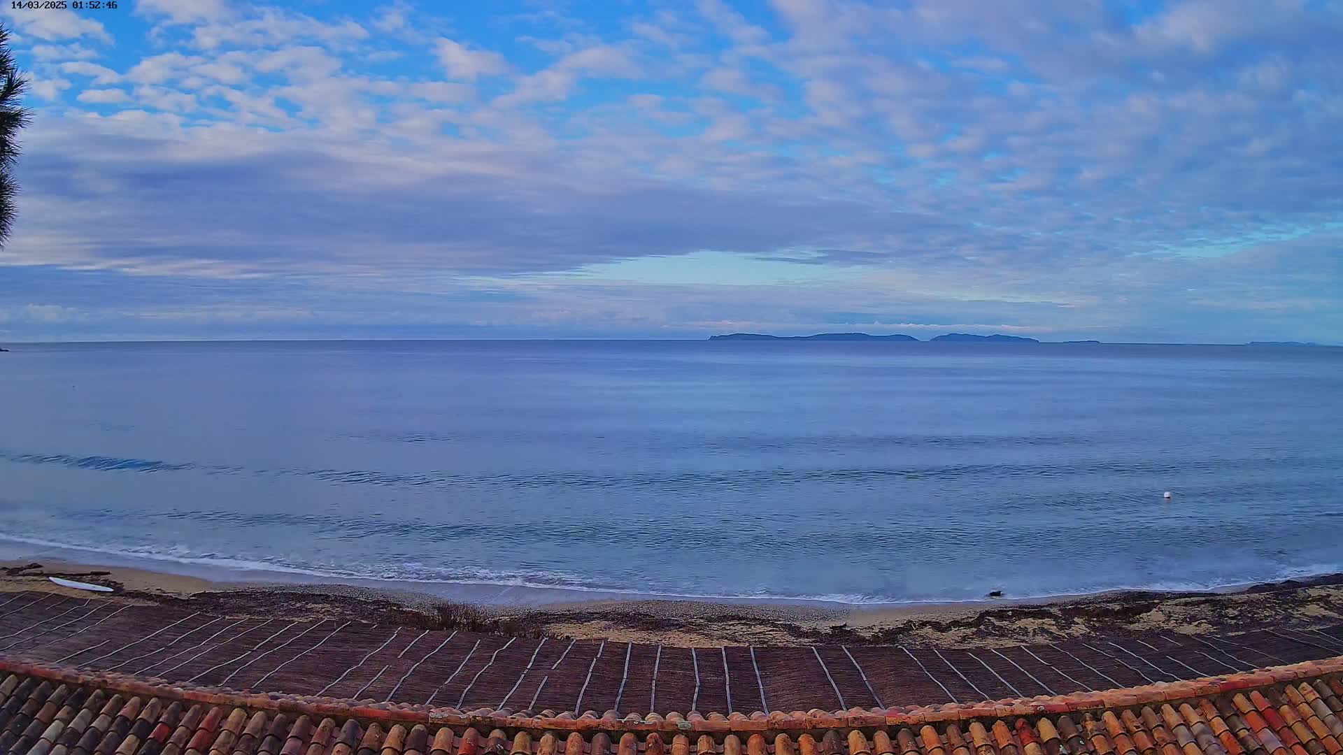 A tranquil ocean scene under a partly cloudy sky shows a sandy beach with a patterned structure in the foreground, and an island visible in the distance.
