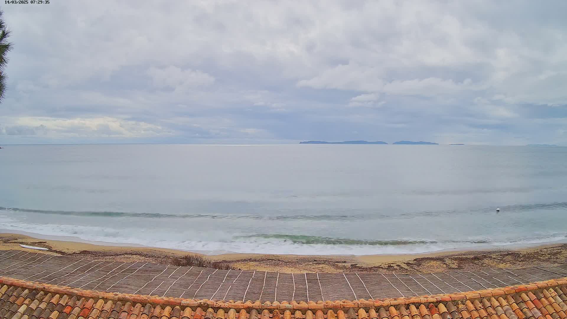 A sandy beach with gentle waves meets a calm sea under a cloudy sky, with distant islands visible on the horizon.