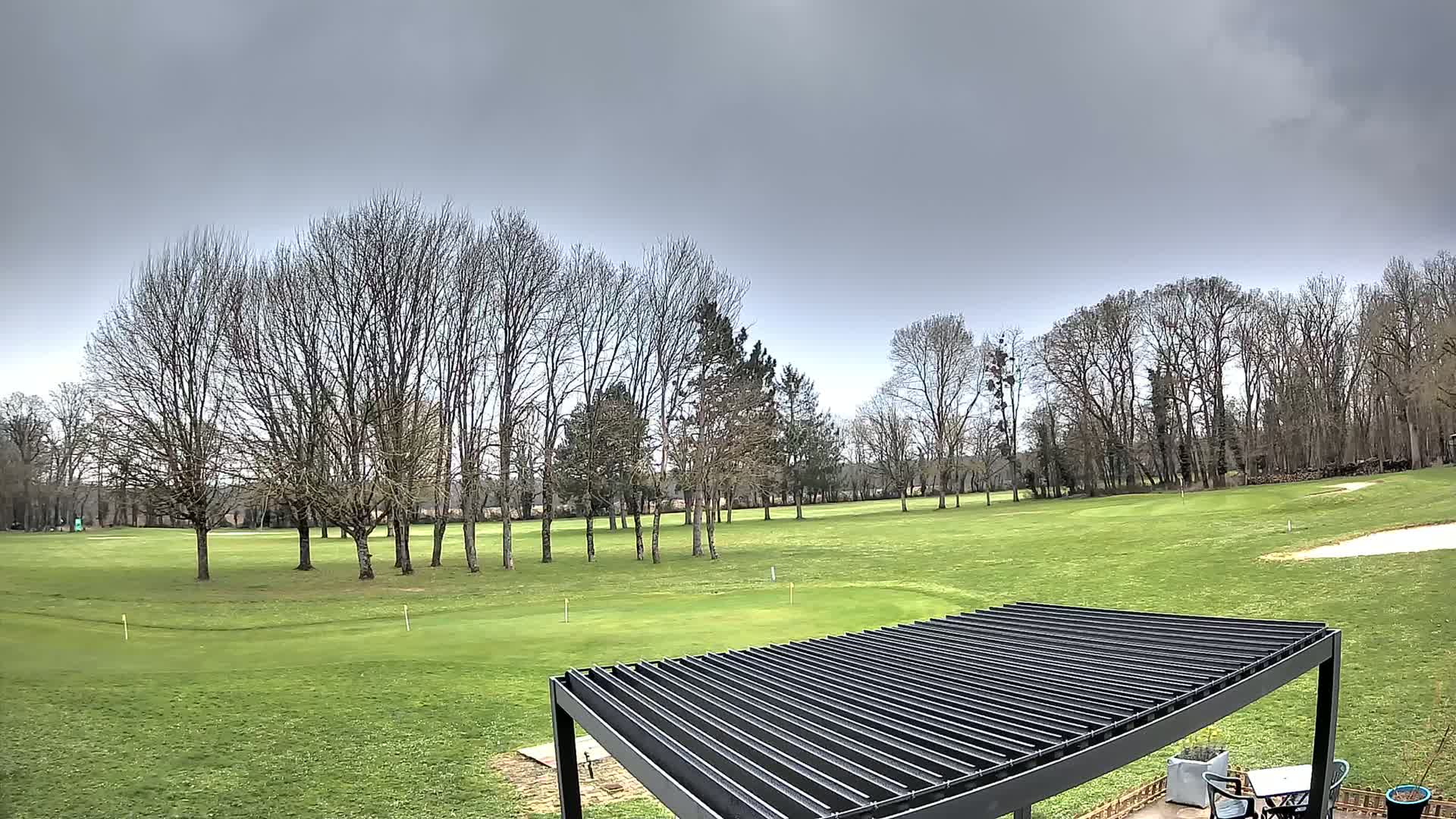 A mostly green golf course is visible under an overcast sky, with bare trees in the foreground and a dark metal pergola in the bottom right corner.