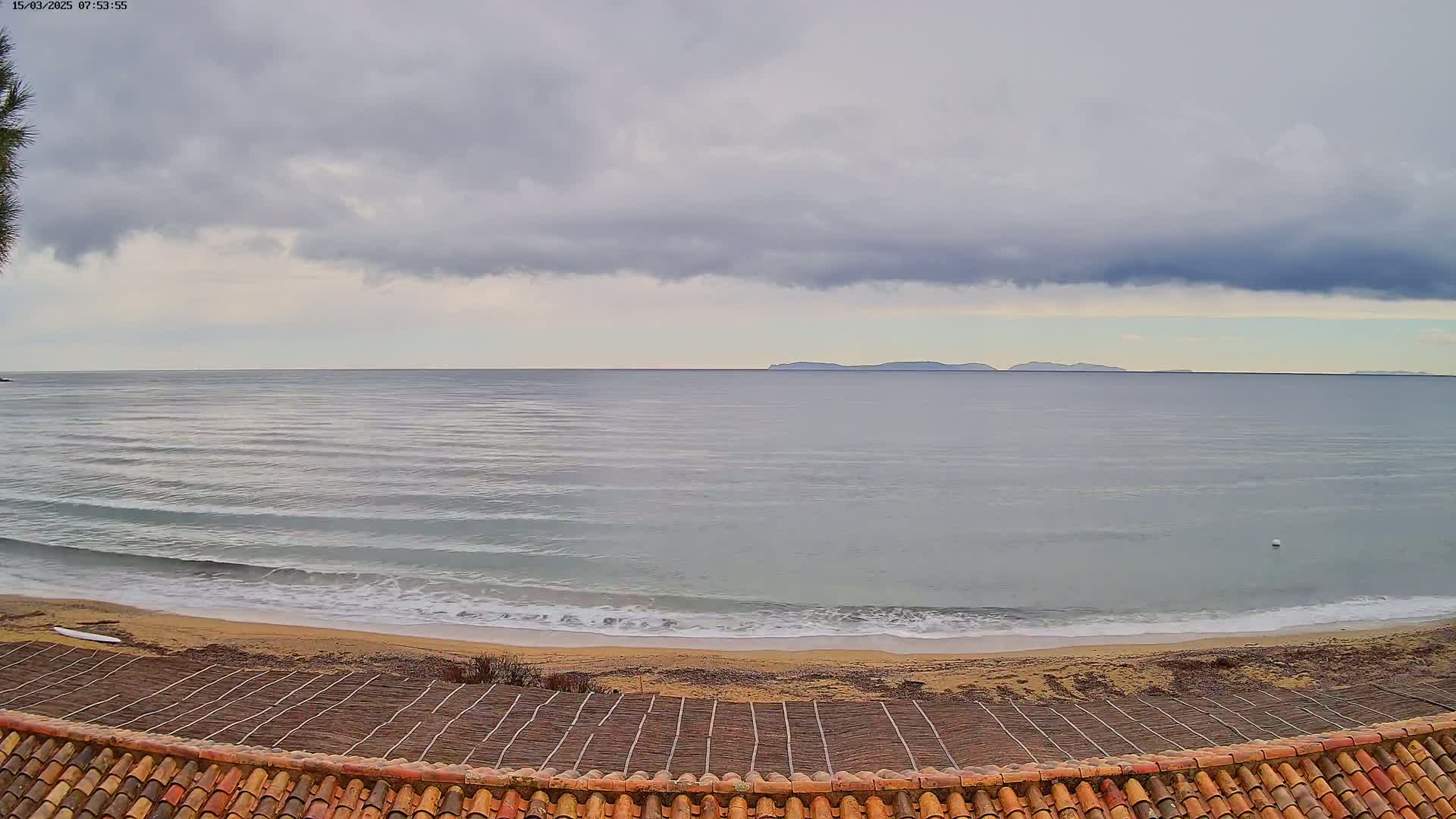 A sandy beach with gentle waves laps the shore under a cloudy sky, with distant islands visible on the horizon.