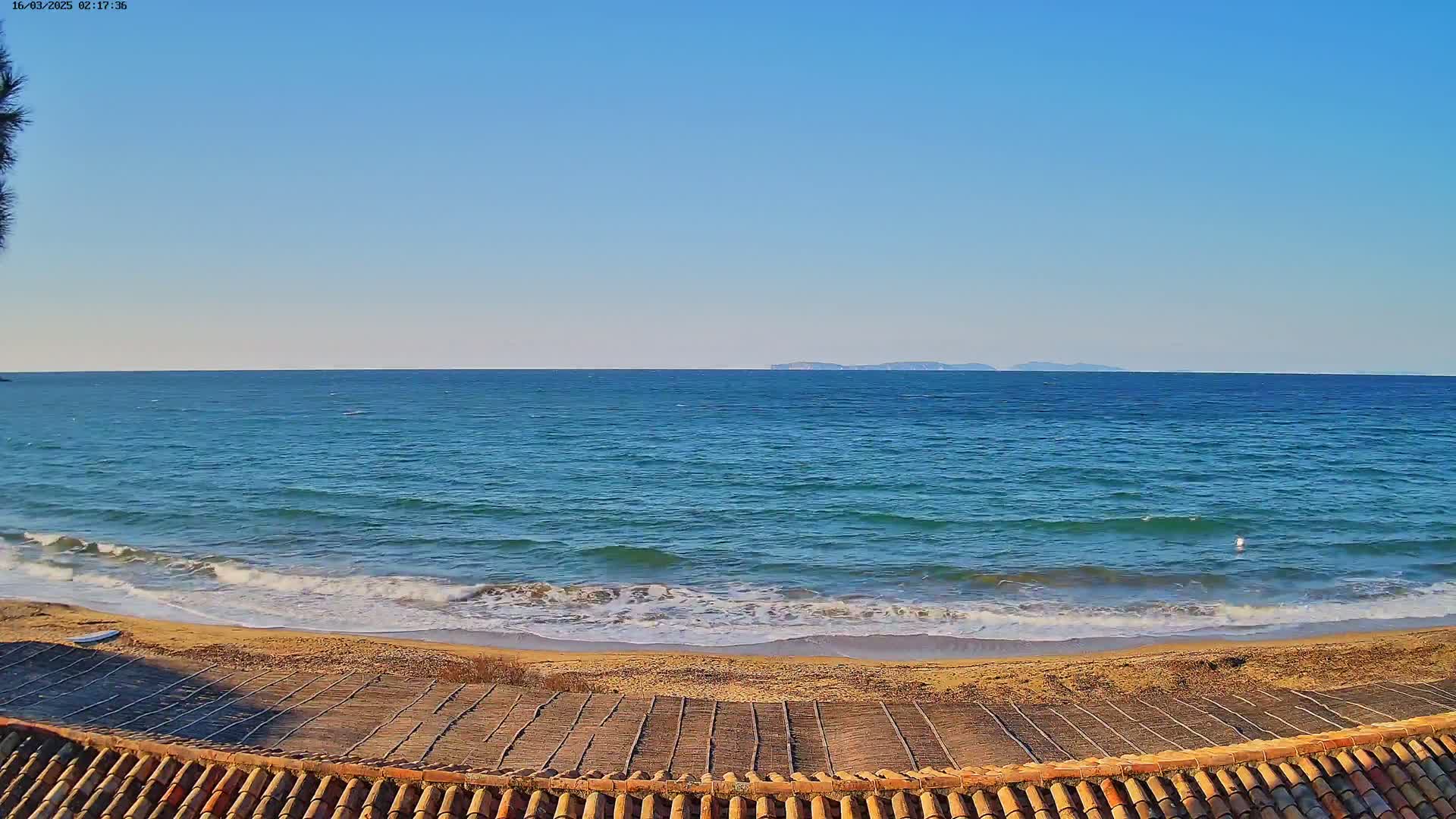 A sandy beach with small waves laps the shore under a clear blue sky, with distant islands visible on the horizon.