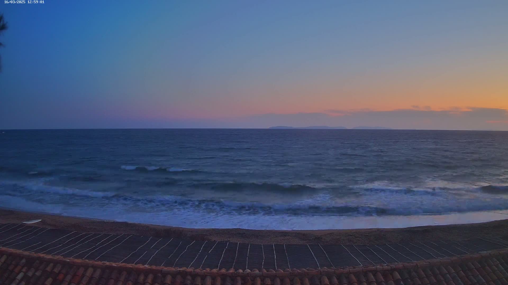 A tranquil ocean scene at sunset shows waves gently lapping a sandy beach with lines of dark material on the sand, viewed from a tiled rooftop.