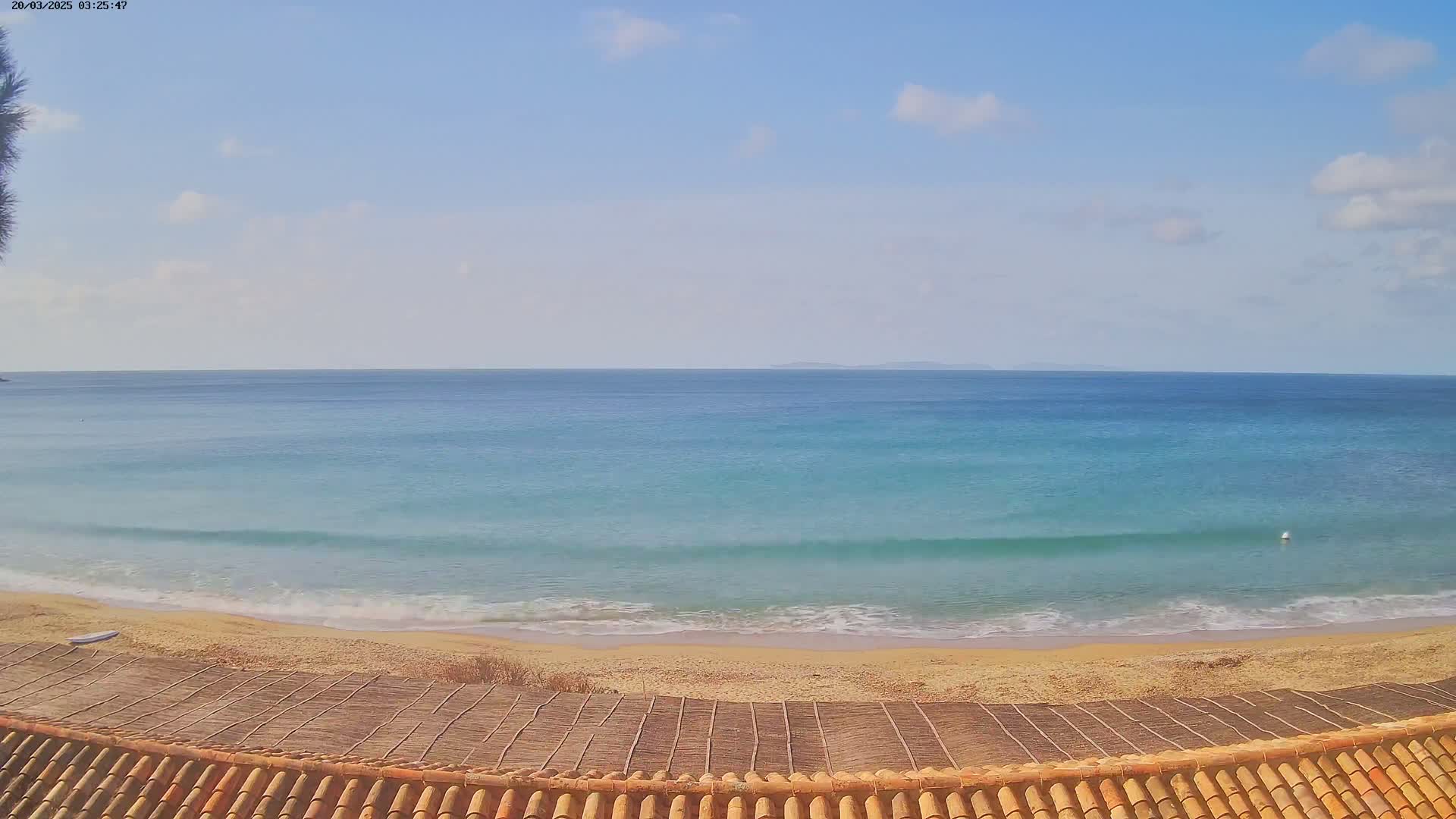 A sandy beach with gentle waves and a calm, partly cloudy sky is viewed from behind a reddish-brown tiled roof.