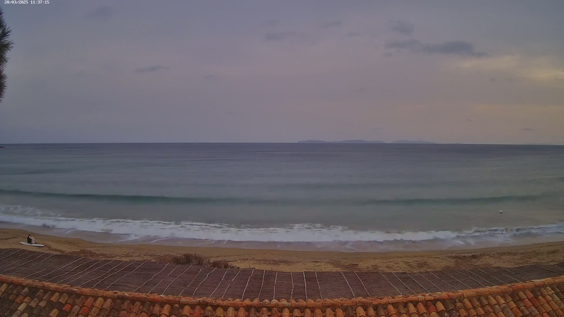 A calm, overcast ocean meets a sandy beach, with a person sitting on a surfboard in the distance, viewed from above a tiled roof.