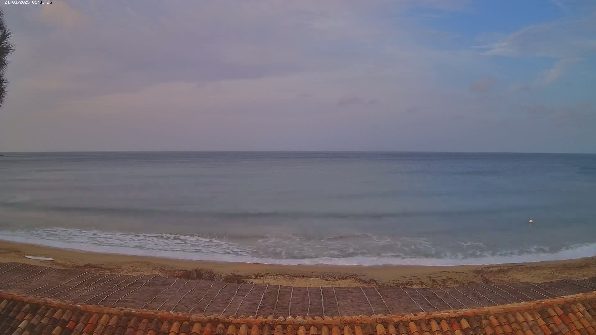 A calm sea with small waves laps a sandy beach under a cloudy, dusky sky, viewed from a tiled roof with a woven matting in the foreground.