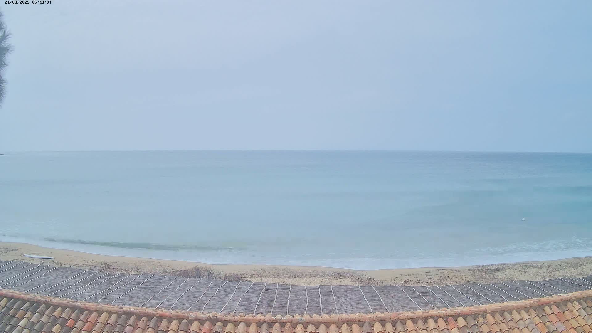 A view from a tiled roof shows a calm, light-blue ocean meeting a sandy beach on an overcast day.