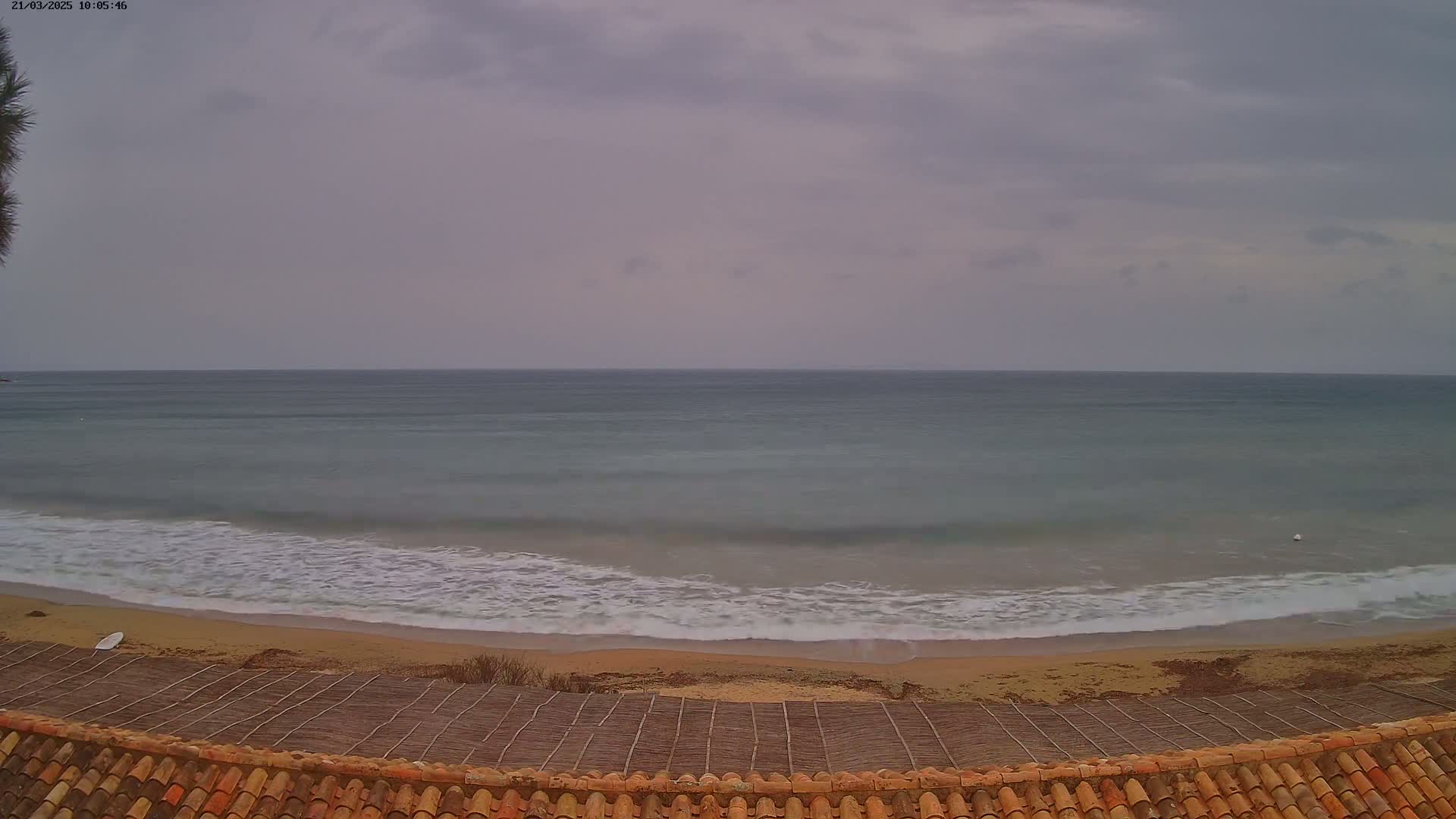 A sandy beach with gentle waves under an overcast sky is viewed from behind a tiled roof.