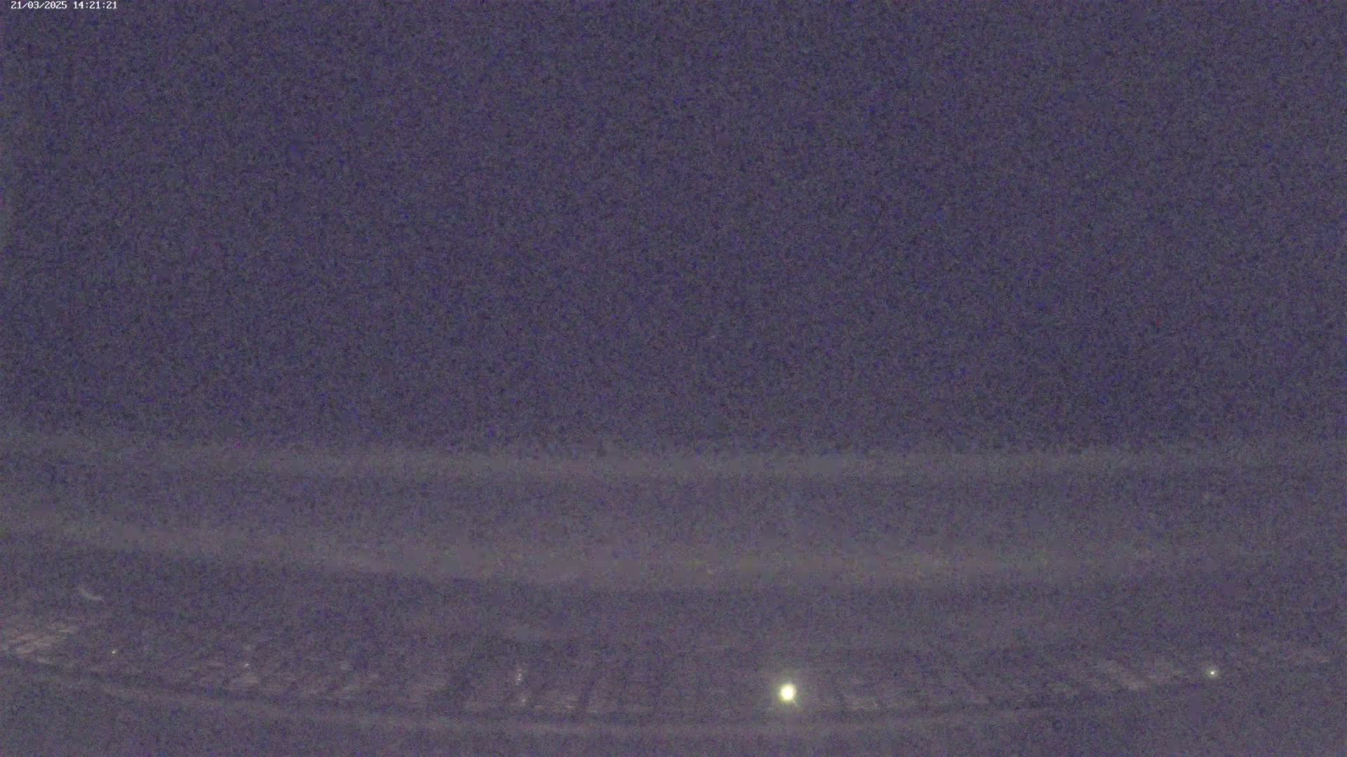 A low-light, nighttime view shows a dark, flat landscape with a few faint lights in the distance under a clear, dark sky.