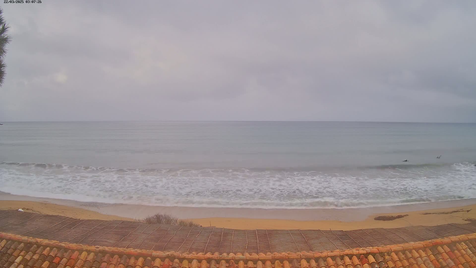 A view from a tiled roof shows a calm sea with small waves lapping a sandy beach under an overcast sky.