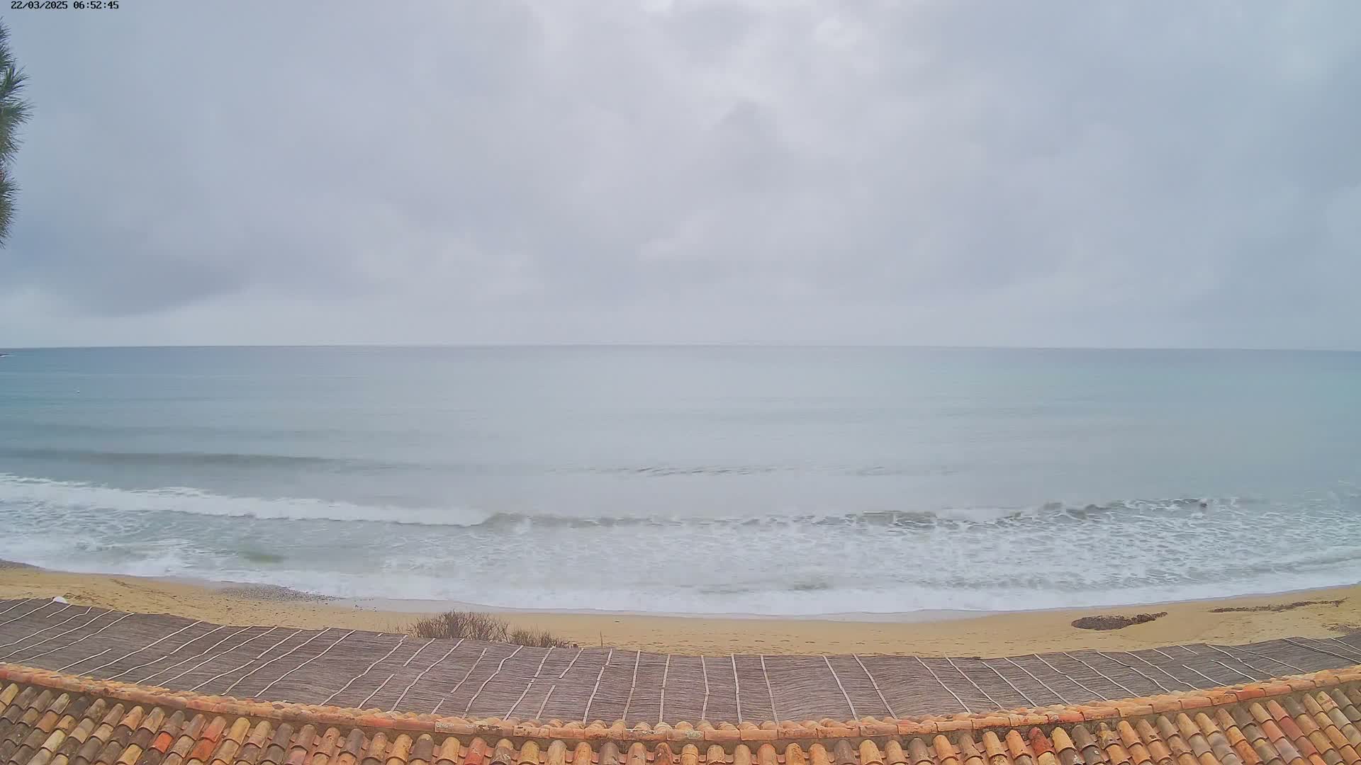 A sandy beach with gentle waves under a cloudy sky is viewed from above a tiled roof.