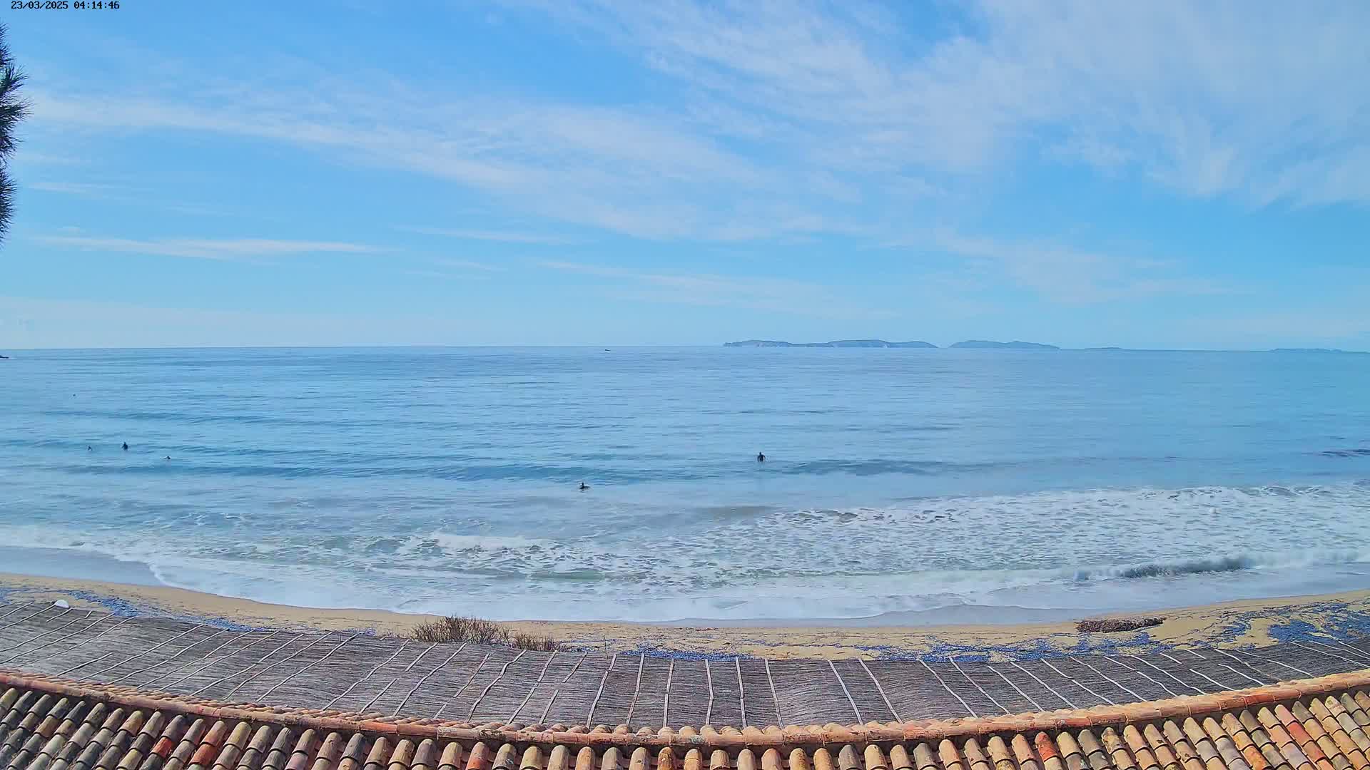 A sandy beach with a few surfers in the ocean under a mostly clear, light blue sky, with islands visible in the distance.