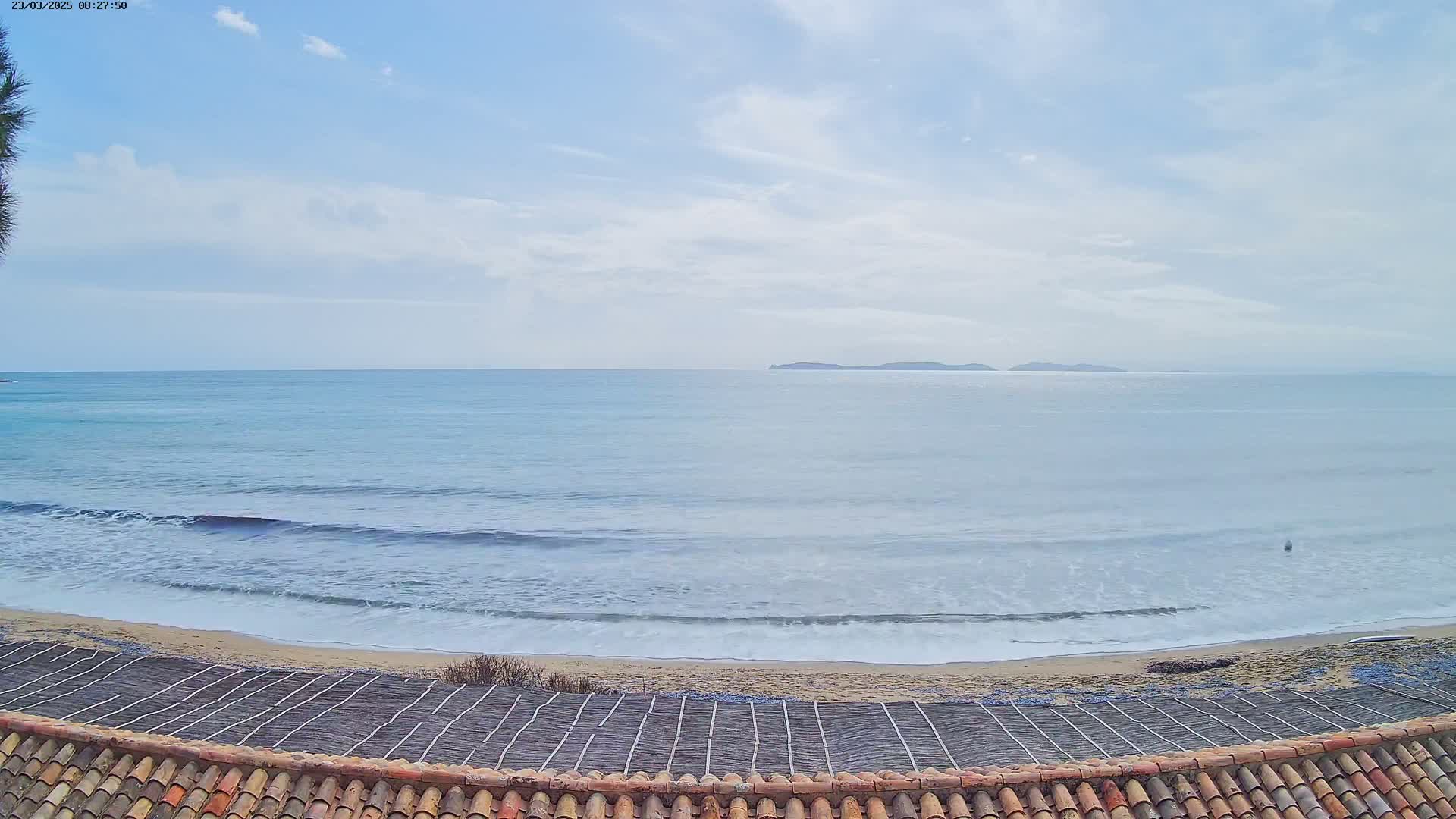 A gently rolling ocean under a mostly clear sky is viewed from above a tiled roof, with a sandy beach and a lattice structure in the foreground.