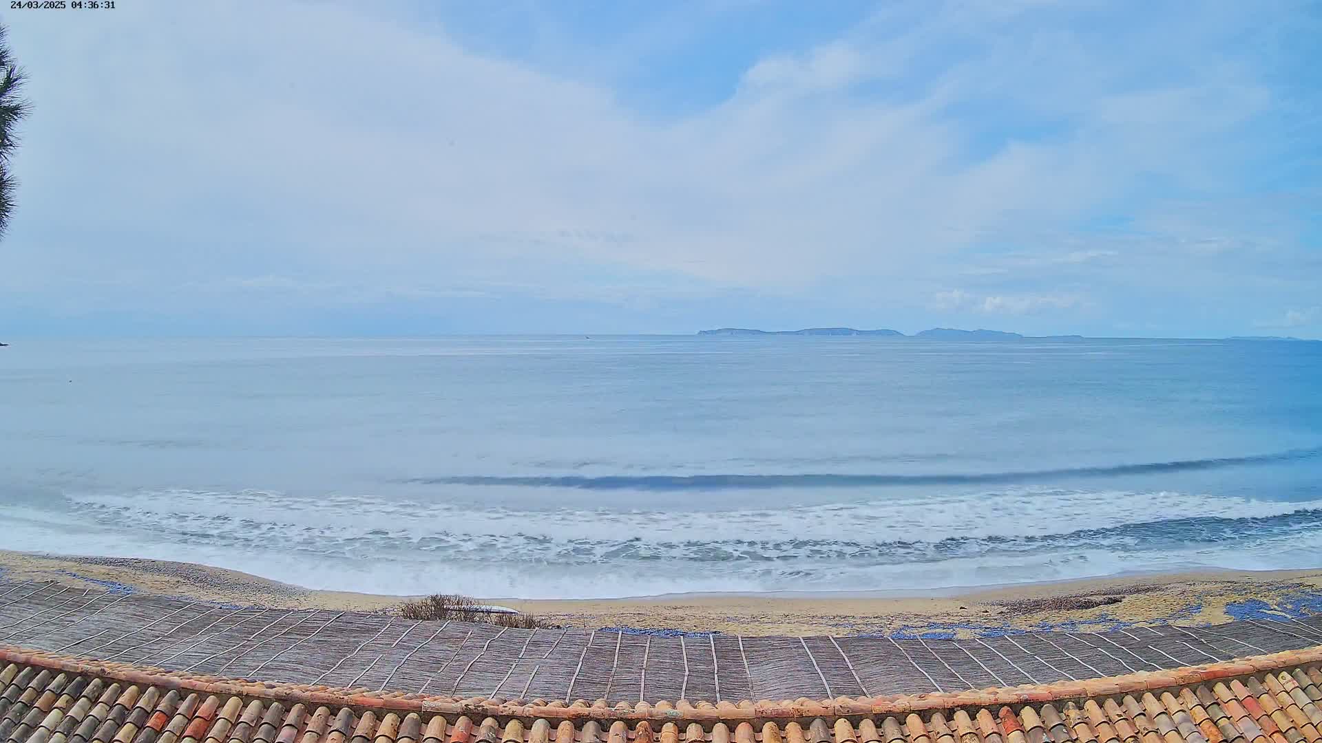 A sandy beach with gentle waves under a mostly cloudy sky, a distant island visible on the horizon.