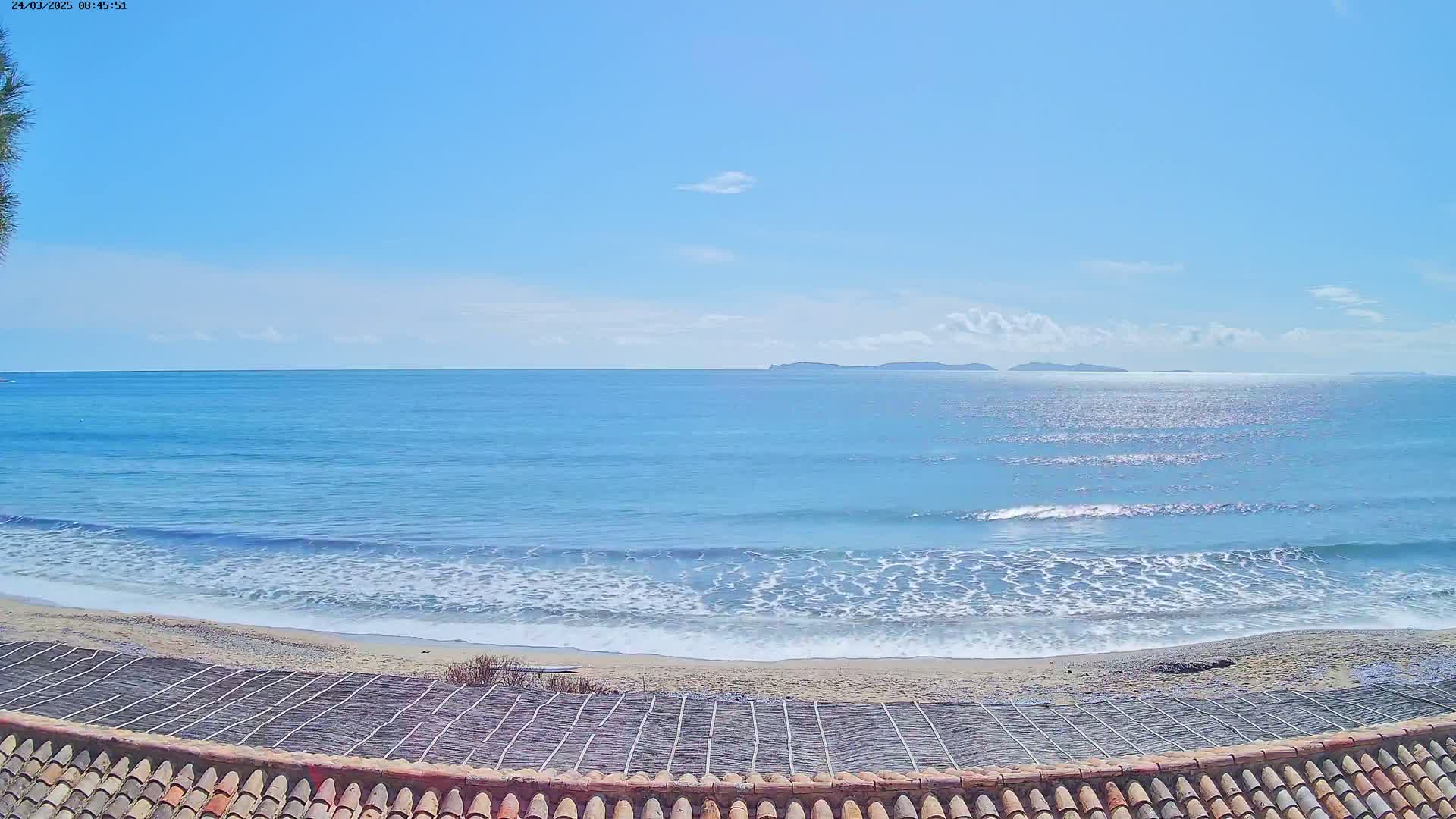 A sandy beach with gentle waves rolls onto the shore under a mostly clear blue sky, with islands visible in the distance.