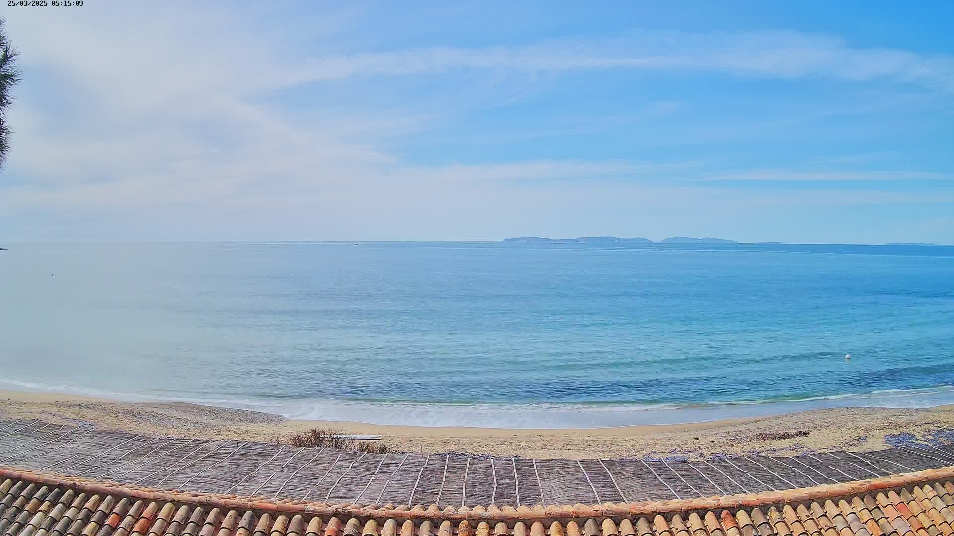 A sandy beach with gentle waves and a calm ocean stretches to a distant island under a mostly clear, bright sky.