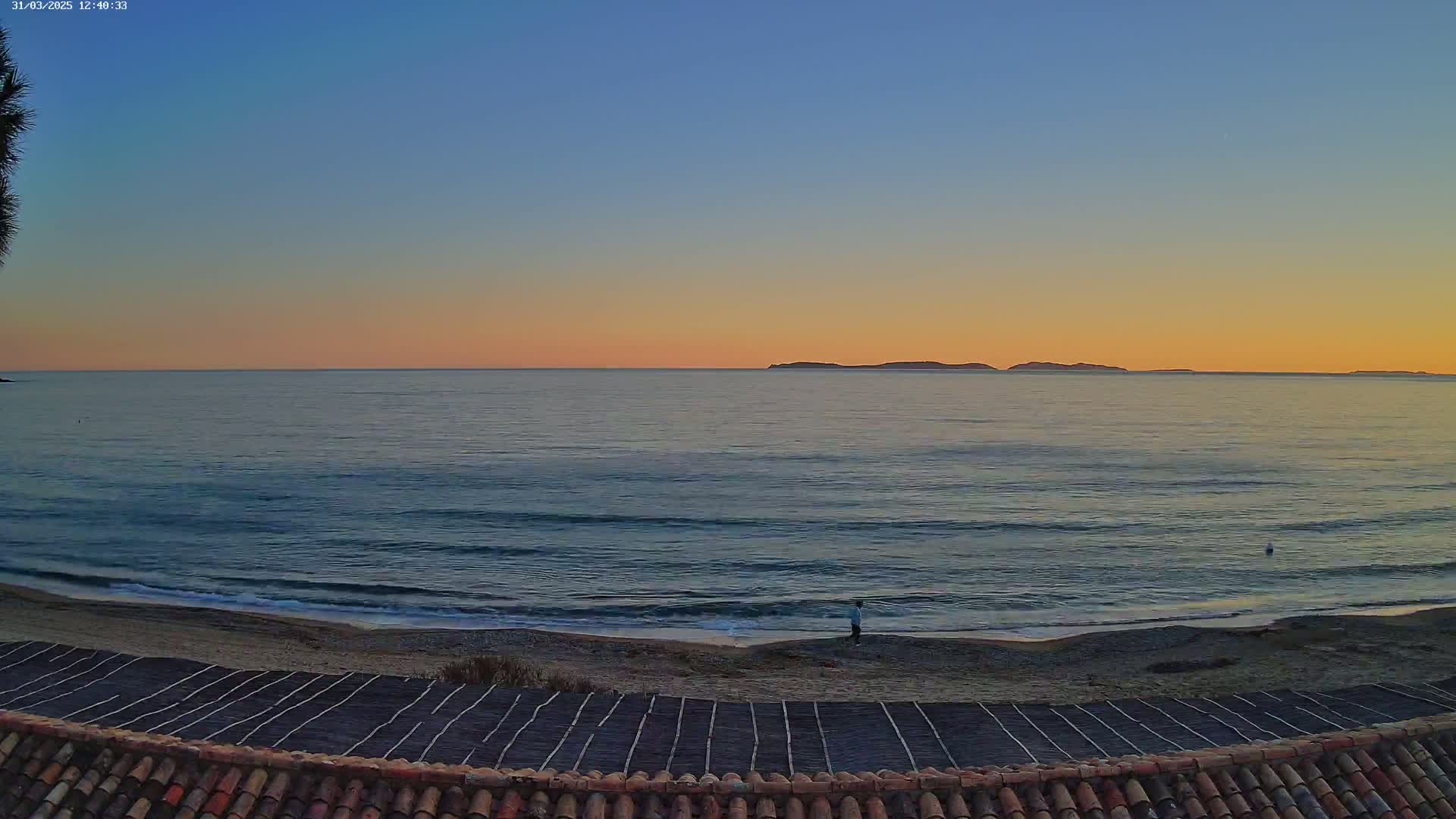 A tranquil ocean scene at sunset shows a calm sea lapping a sandy beach, with a lone figure standing near the water's edge and distant islands on the horizon.