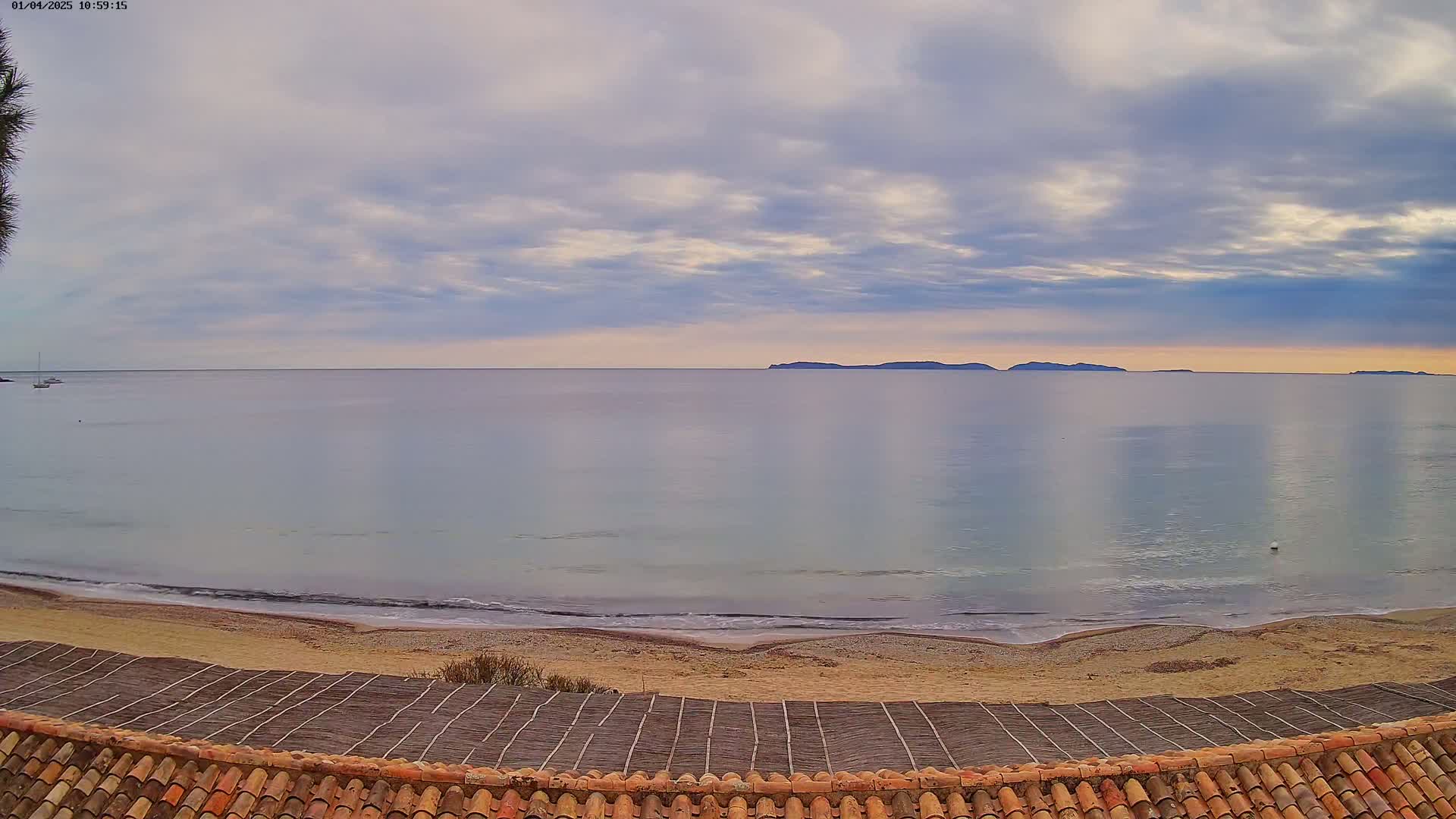 A calm, light-grey sea with a sandy beach and distant islands is seen from above a tiled roof under a partly cloudy sky.
