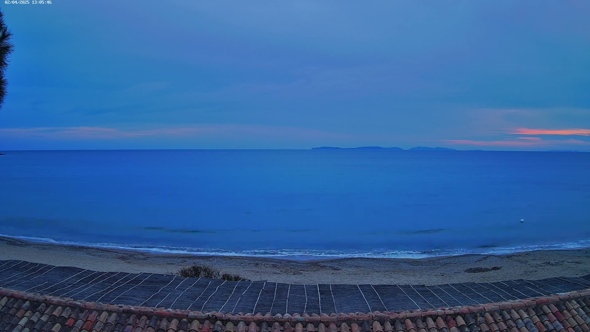 A tranquil beach scene at dusk shows a calm sea meeting a sandy shore, with a distant island and a tiled roof in the foreground.